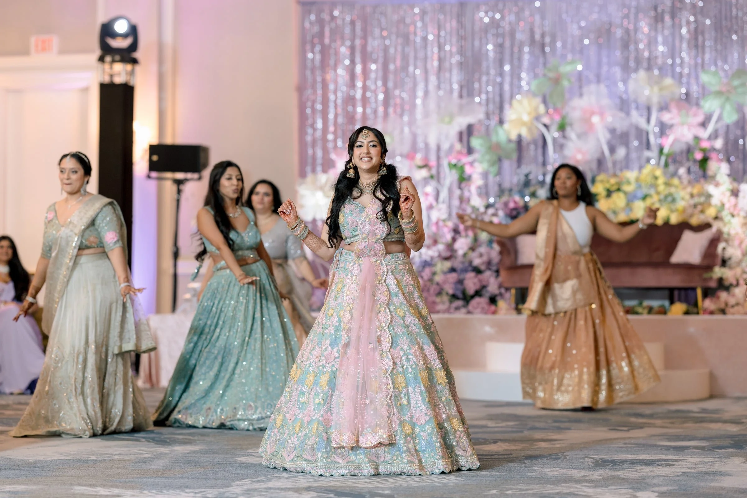 Women dressed in traditional Indian wedding attire dancing at a celebration, with floral decorations and shimmering backdrop.