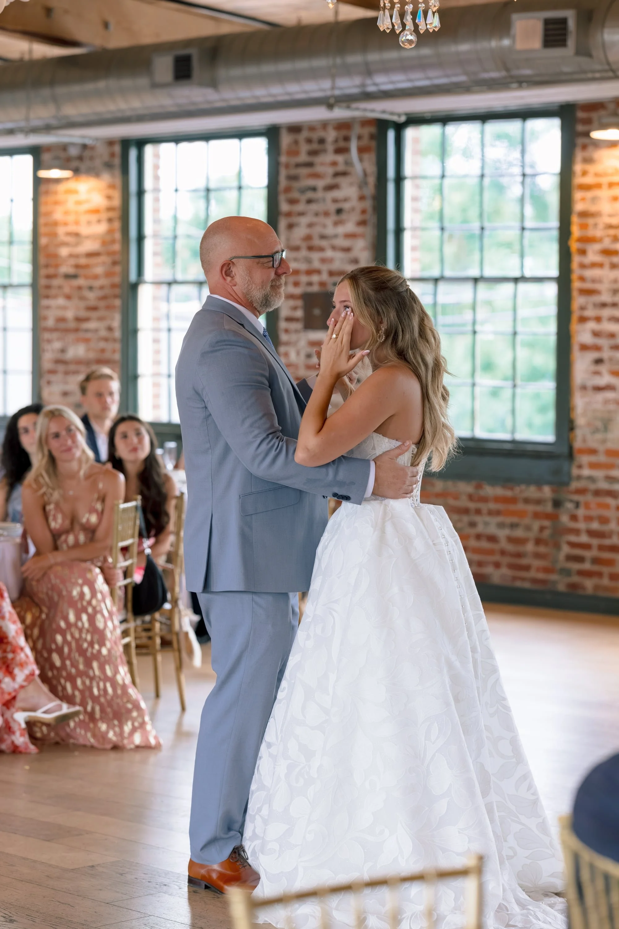 A bride and groom sharing a dance during their wedding reception, with guests watching in the background inside a venue with brick walls and large windows.