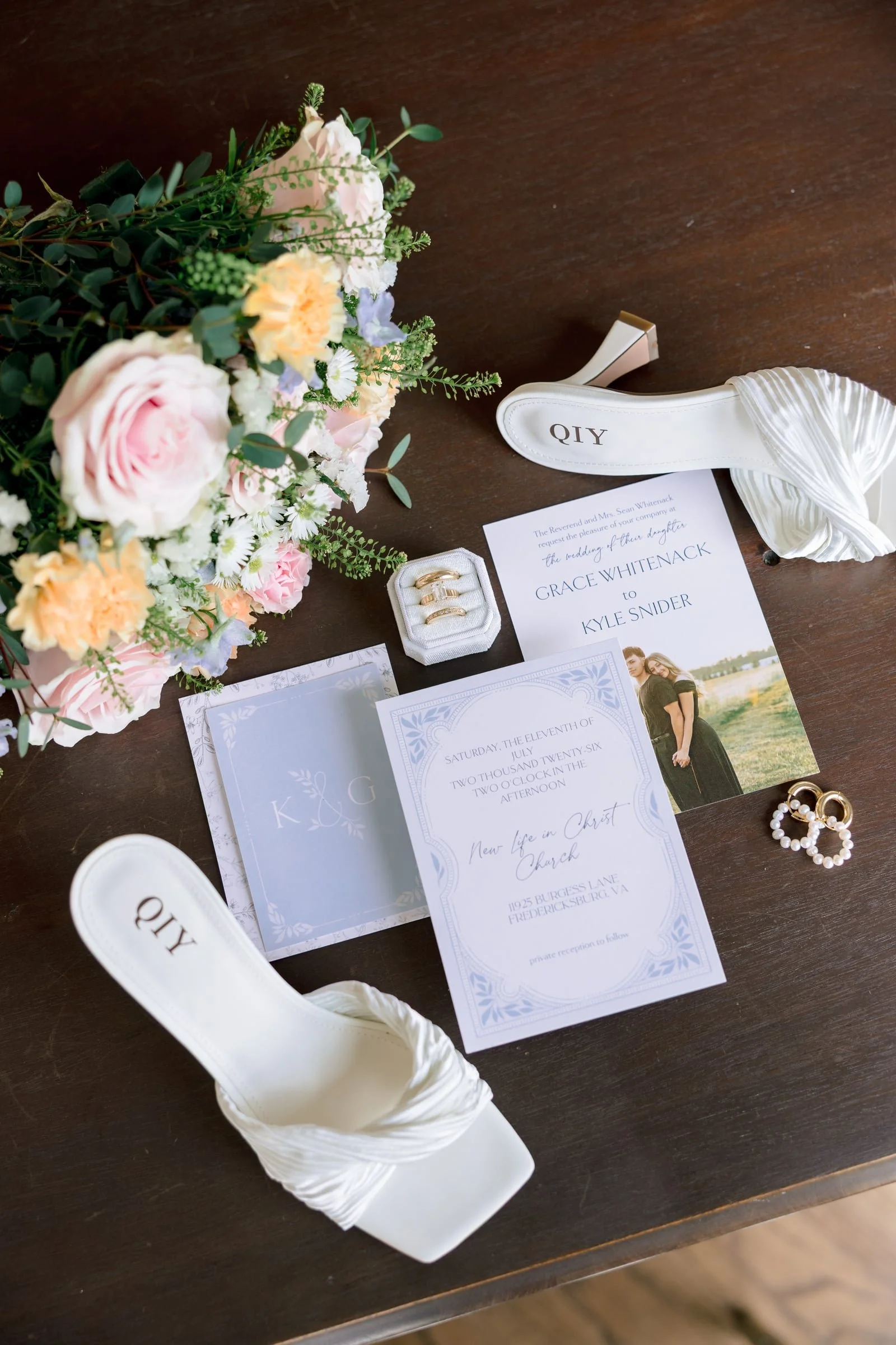 Wedding invitation, bouquet of flowers, rings, and white high-heeled shoes on a wooden table.