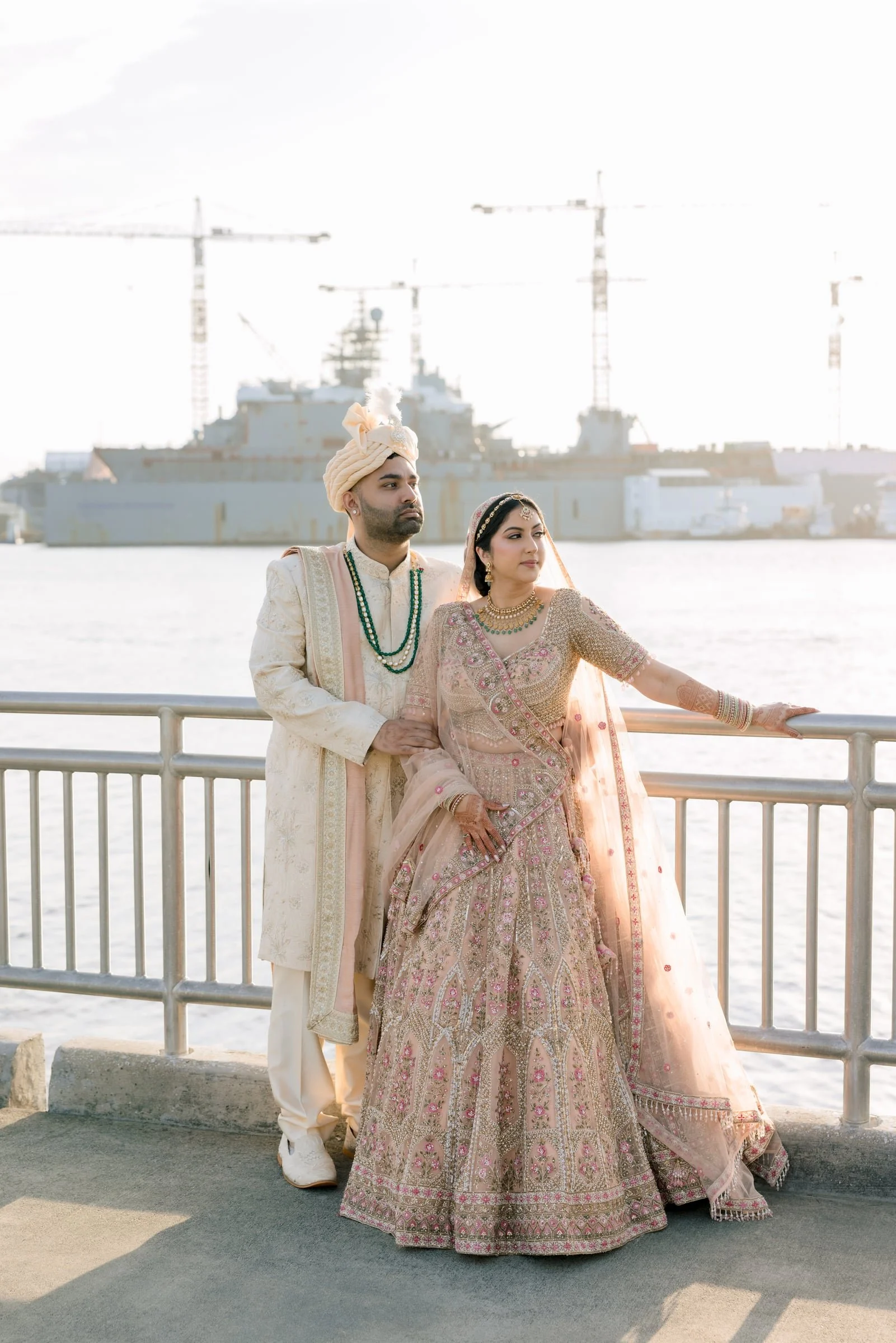 An Indian couple dressed in traditional wedding attire, standing beside a waterfront railing with large ships and construction cranes in the background, during sunset or sunrise.