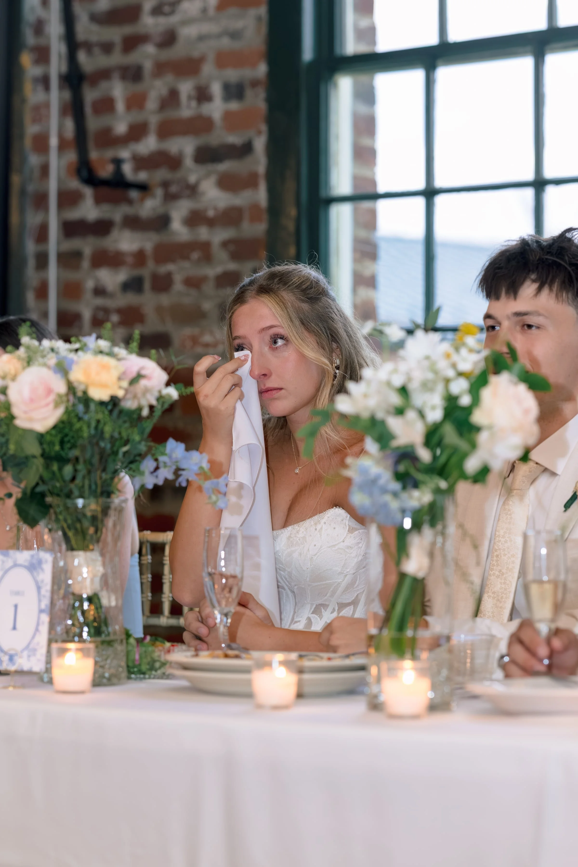 A bride wiping tears while sitting at a decorated wedding table with floral centerpieces and candles, alongside a groom and guests, during a wedding celebration in a rustic venue with brick walls and large window.