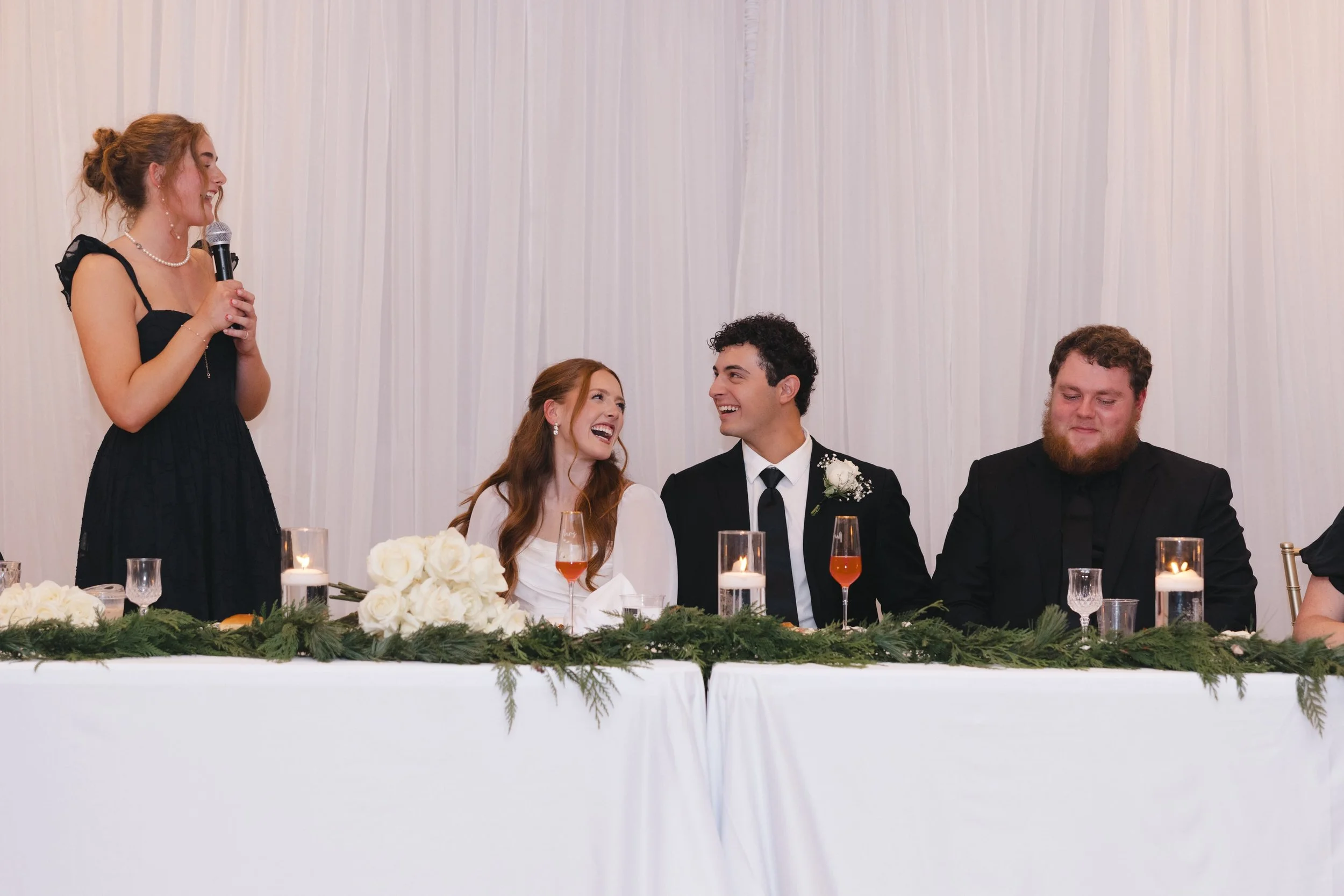 A woman in a black dress holding a microphone, giving a speech at a wedding reception. Three people are seated at a decorated table with white flowers, candles, and drinks, smiling and listening.