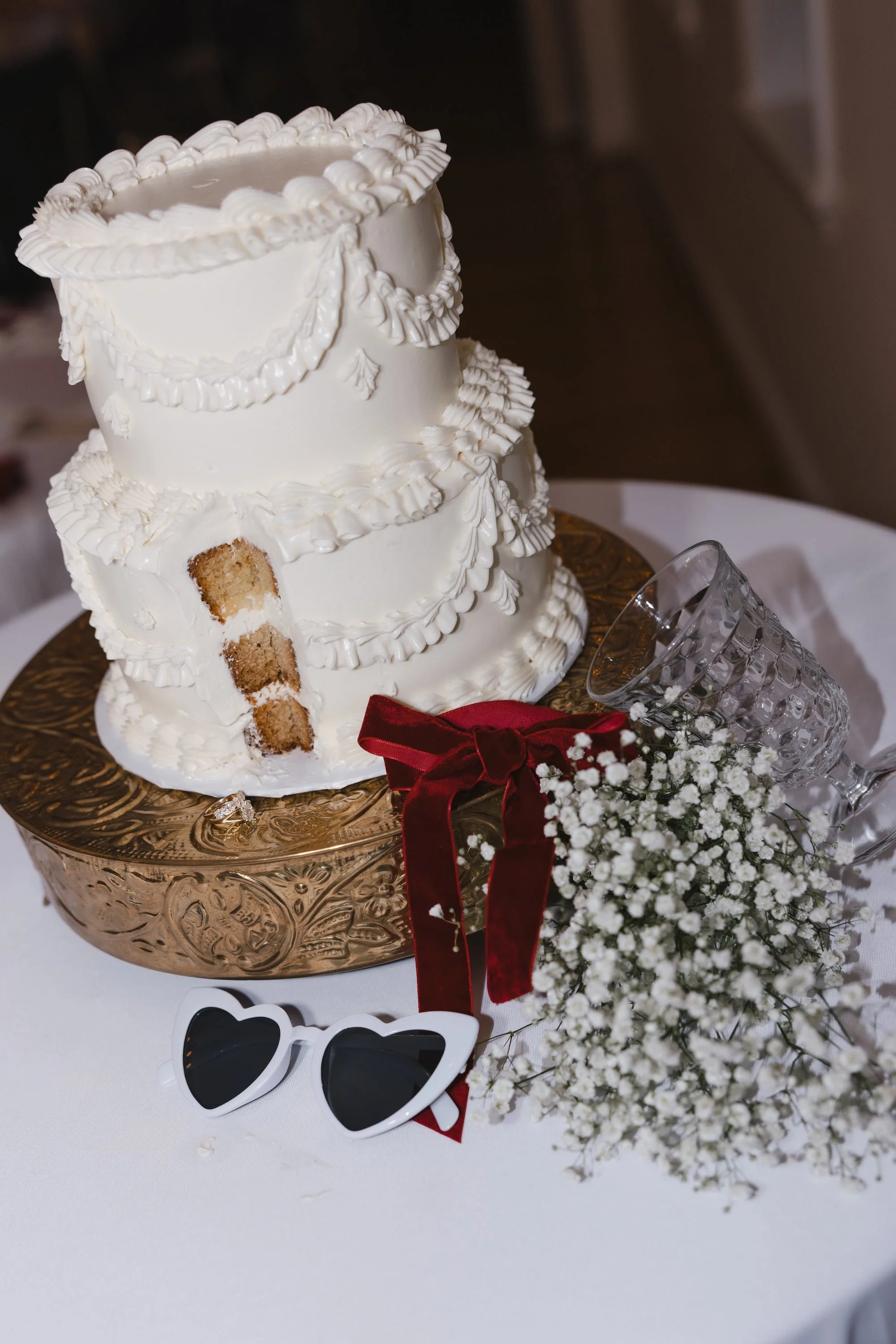 A white, multi-tier wedding cake with decorative piping and a partially eaten slice placed on a decorative gold cake stand. Beside the cake, there are heart-shaped sunglasses, a small bouquet of white baby's breath flowers with a red ribbon, and a cl