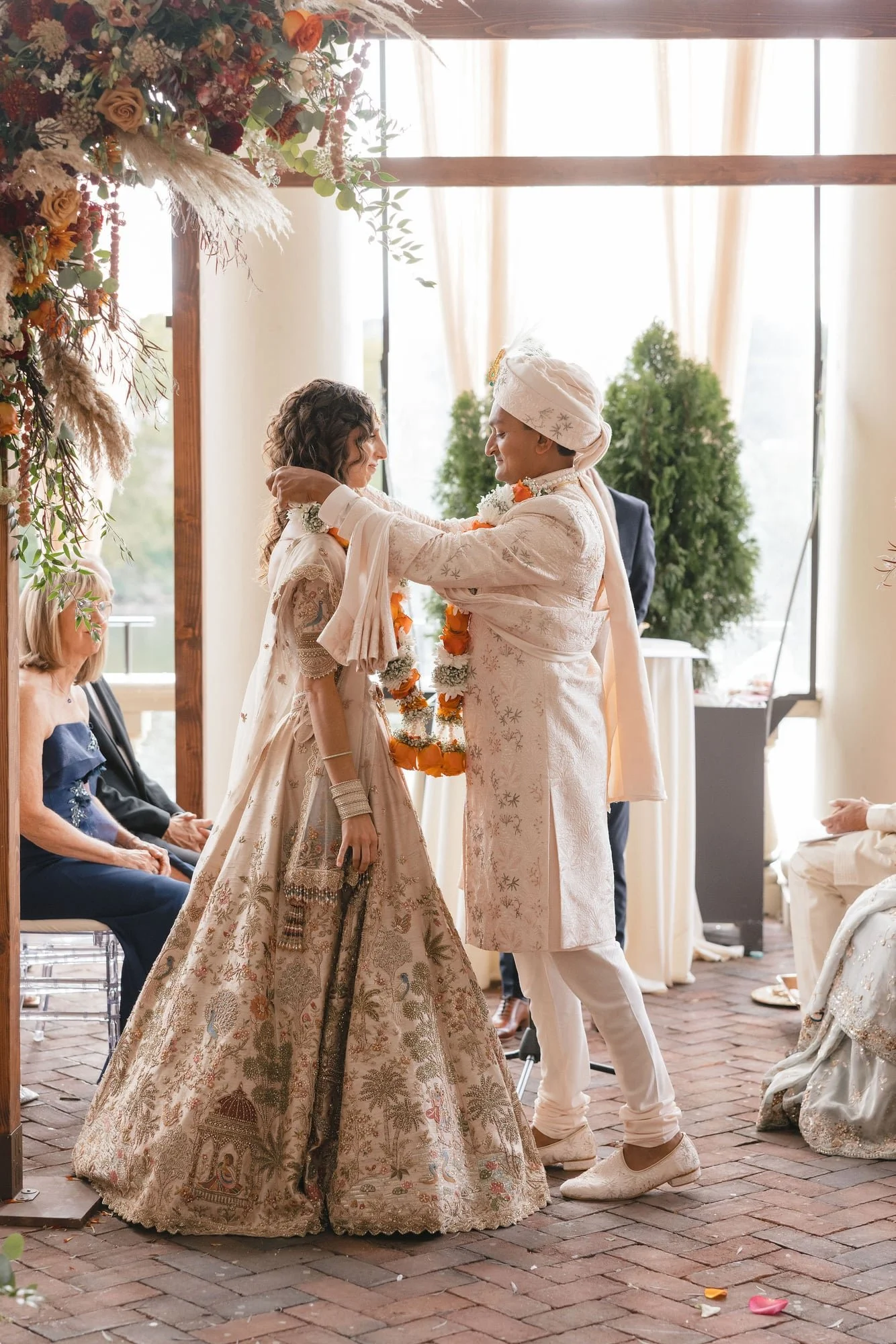 Two women are celebrating a wedding, standing close with their foreheads touching. One is wearing a traditional brown and gold embroidered dress, and the other is in a cream-colored jacket and turban with floral decorations. Guests are seated nearby,