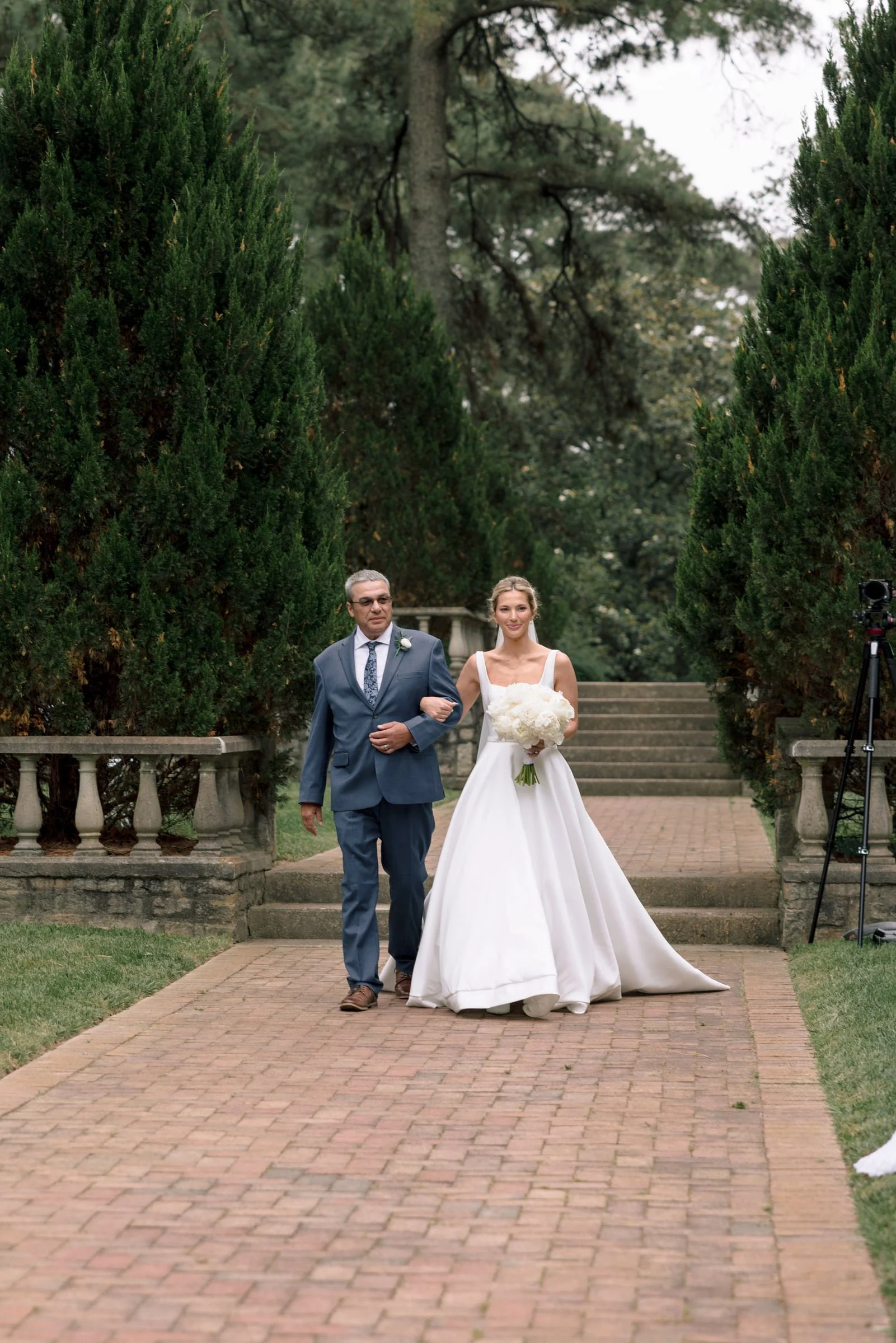 A bride walking down the aisle with her father during a wedding ceremony outdoors. The bride is wearing a white wedding gown and holding a bouquet of white flowers. The father is wearing a grey suit with a patterned tie.