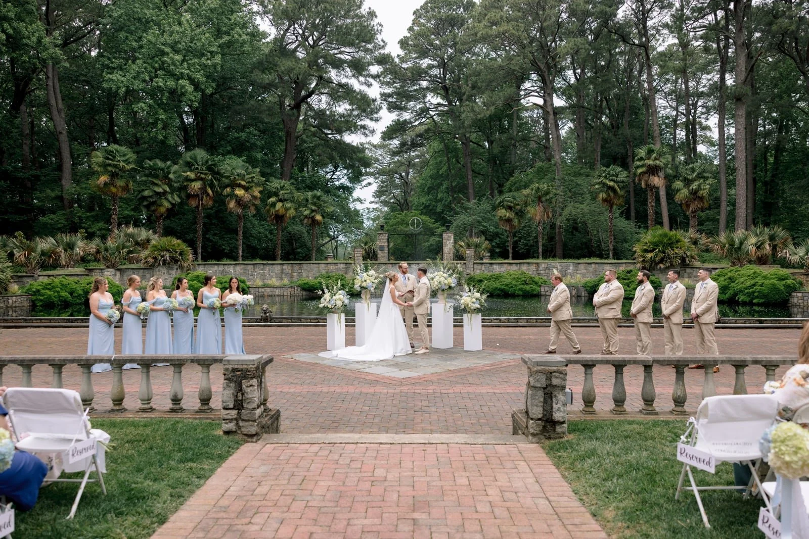 A wedding ceremony taking place outdoors in front of a pond and lush green trees. The bride and groom are standing in the center, holding hands. The bride wears a white wedding dress, and the groom wears a beige suit. Bridesmaids in light blue dresse