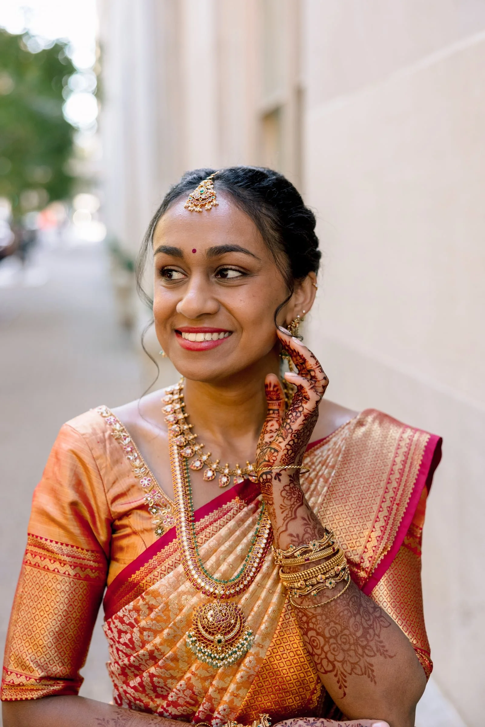 A woman dressed in traditional Indian attire, wearing a gold and pink saree, ornate jewelry including necklaces, earrings, and bangles, with henna designs on her hand, smiling and touching her earring.