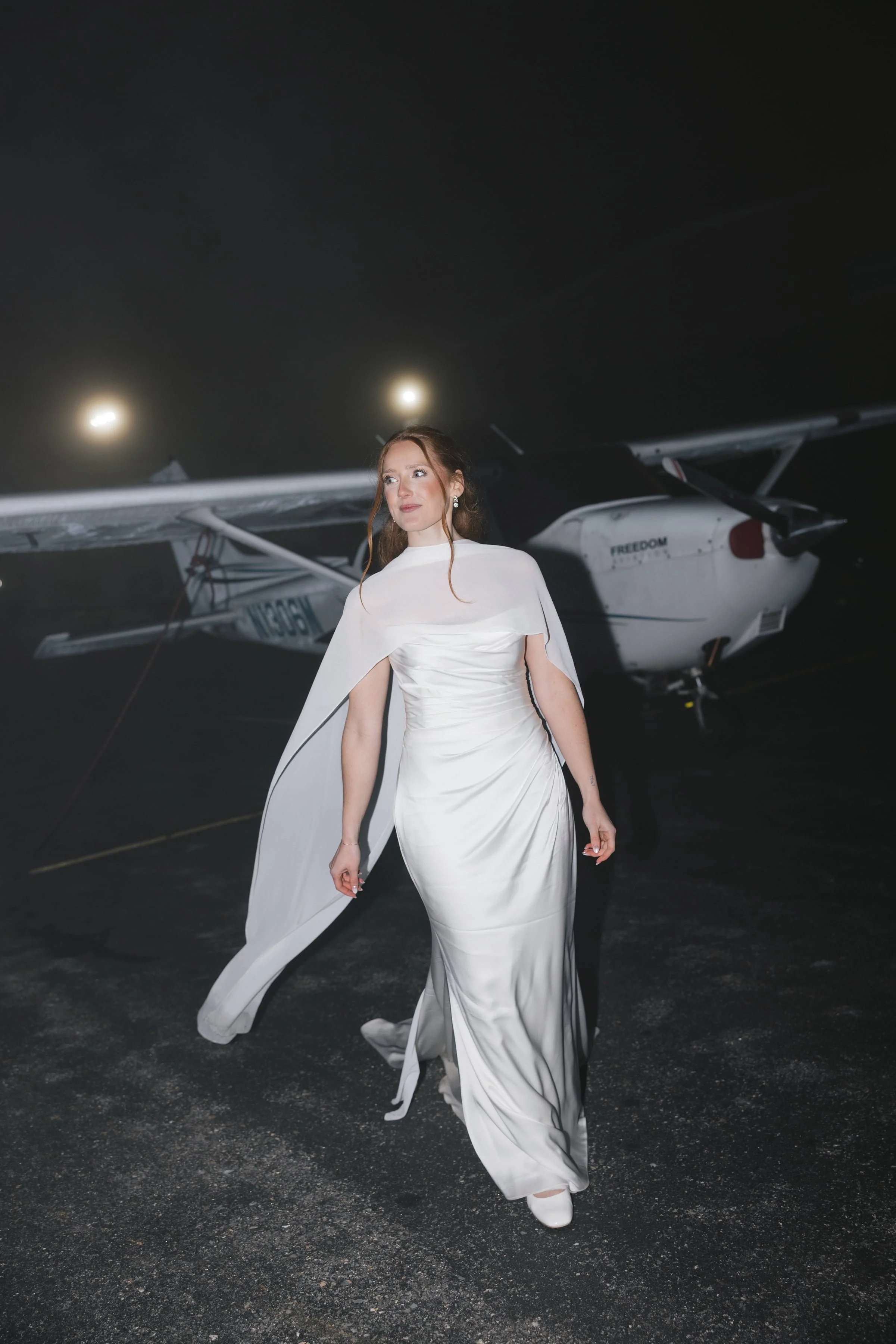 A woman in a white satin gown with a cape and white shoes stands outdoors at night, with a small aircraft in the background.