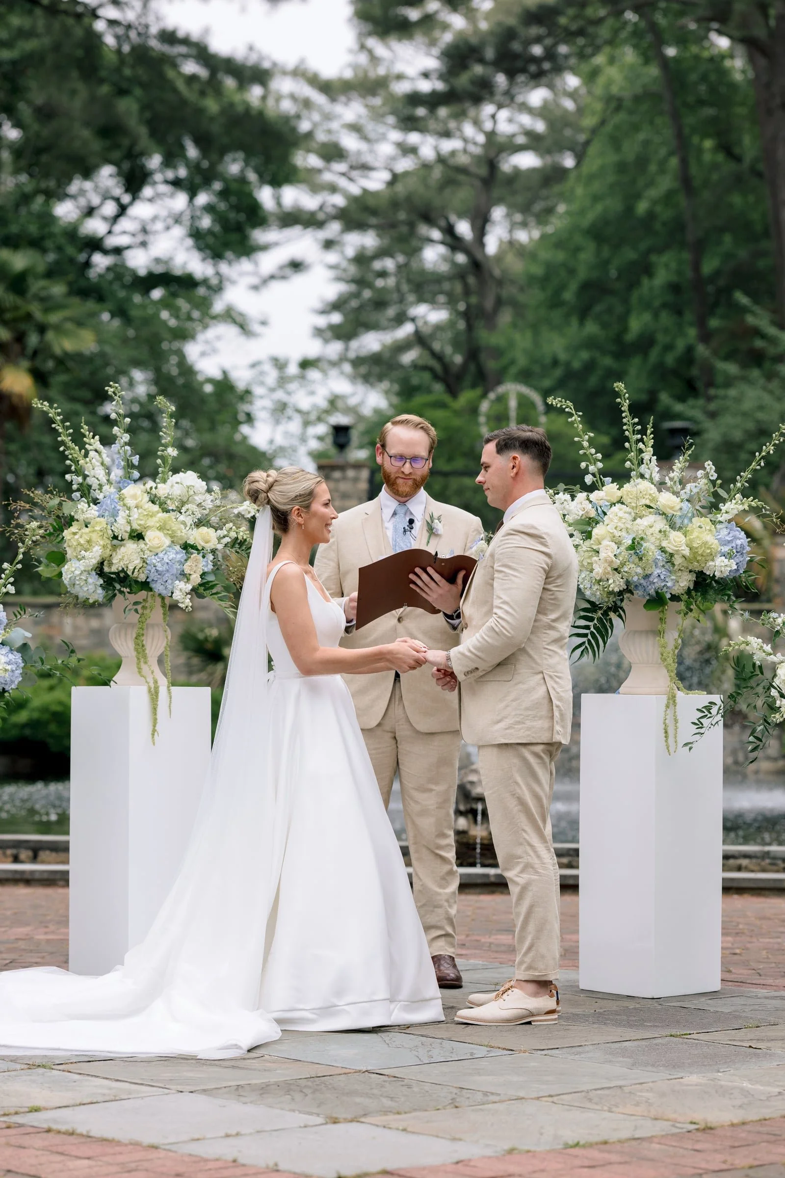 A wedding ceremony outdoors with a bride and groom holding hands and facing each other, with a officiant standing behind them. The bride is in a white wedding dress with a veil, and the groom is in a tan suit. The ceremony is decorated with large flo