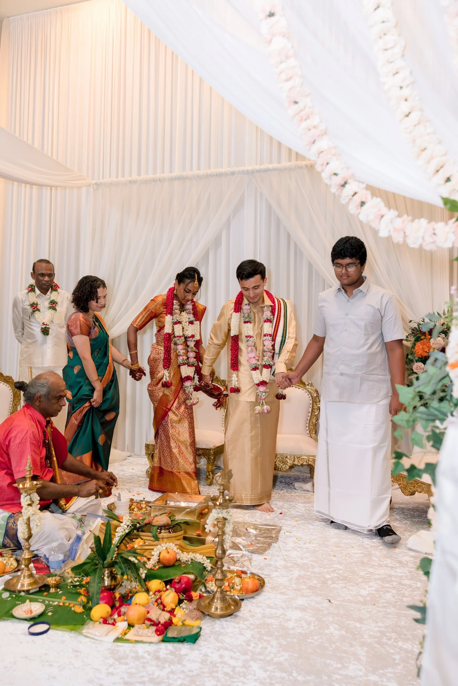 A traditional Indian wedding ceremony with the bride and groom standing on a decorated stage, surrounded by family members, with offerings, fruits, flowers, and wedding items in the foreground.