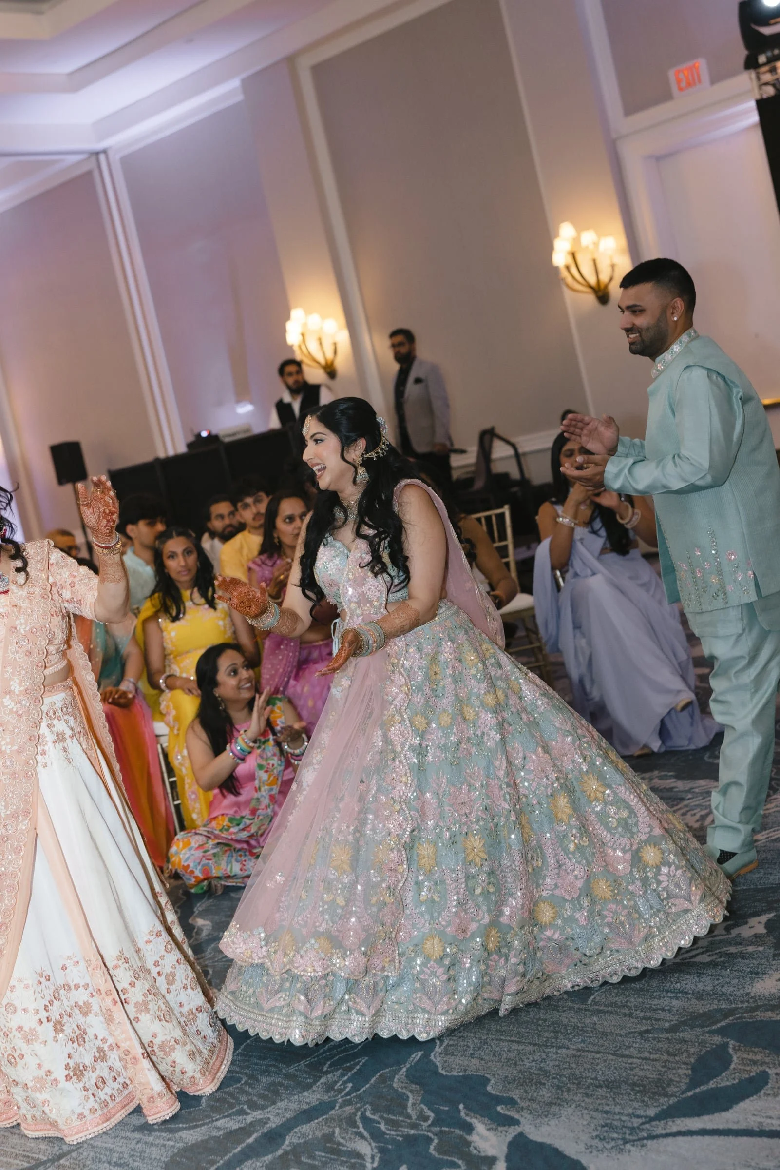 A group of people at a celebration, with a woman in a pink and gold dress dancing in the foreground. The woman is wearing traditional Indian attire with bangles, earrings, and a headpiece. Other guests are watching and smiling, with some sitting and 