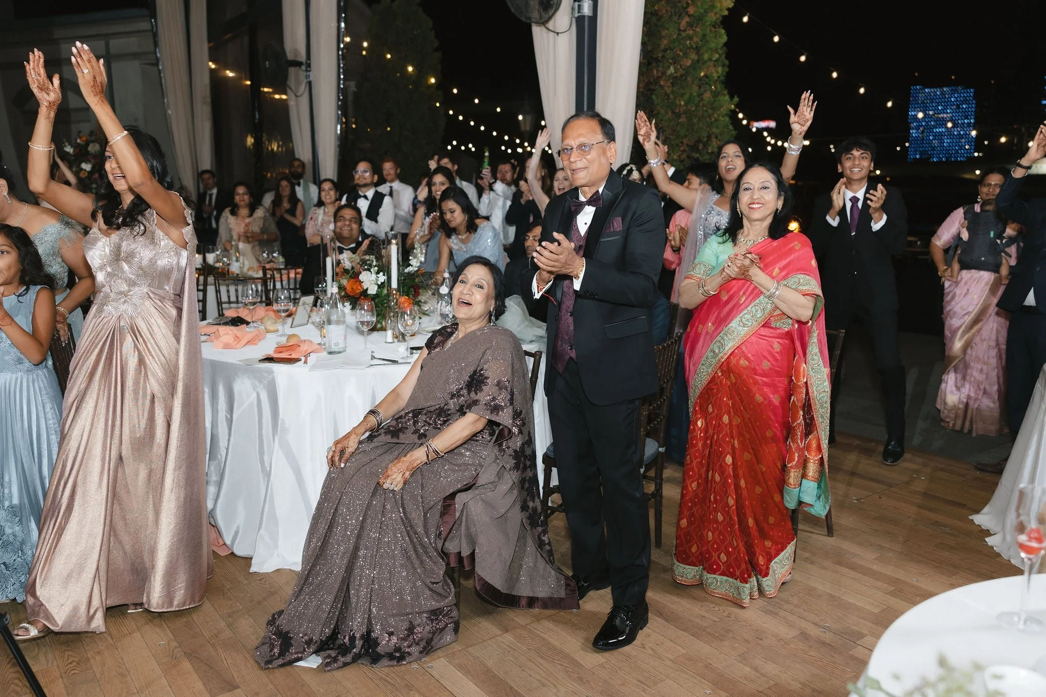 A group of people celebrating at a nighttime event. Several guests are dancing and smiling, with some clapping. The scene is decorated with string lights, and a table with floral arrangements and glasses is visible in the foreground. The guests are w