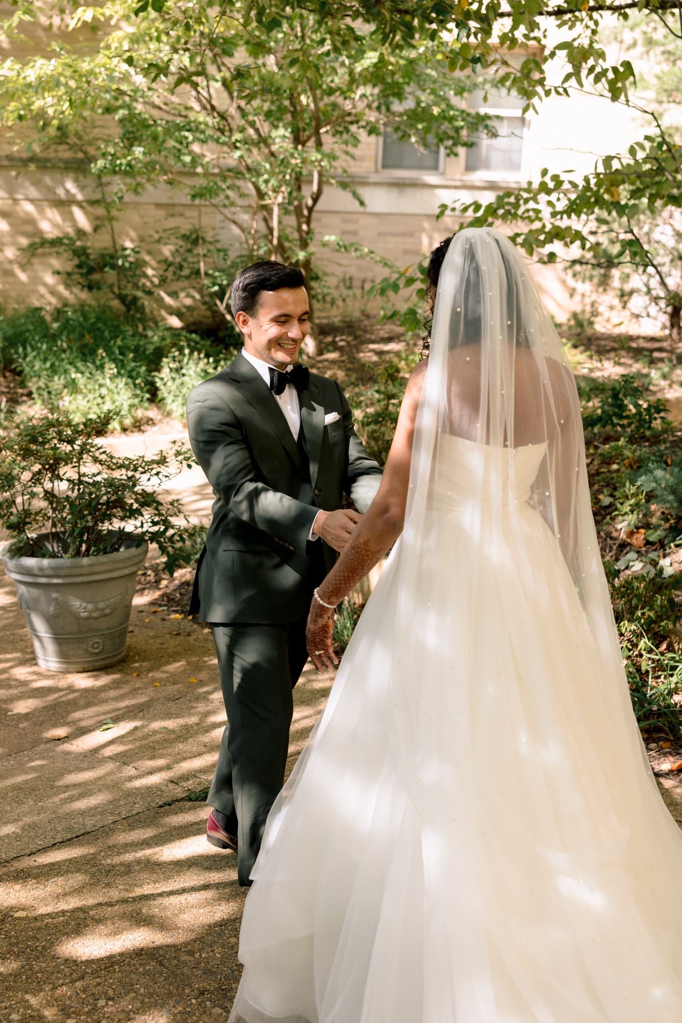 A bride and groom holding hands outdoors. The groom is wearing a black tuxedo with a bowtie, smiling, and looking at the bride. The bride is wearing a white wedding gown and veil, facing the groom. The setting is a garden with trees, bushes, and a bu