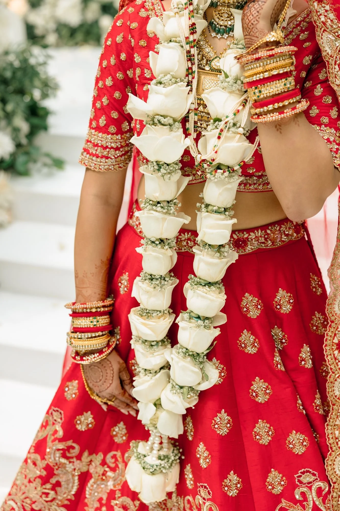 A woman dressed in a traditional red and gold embroidered Indian bridal outfit, adorned with multiple colorful bangles, wearing layered jewelry, and a long garland made of white flowers and white roses.