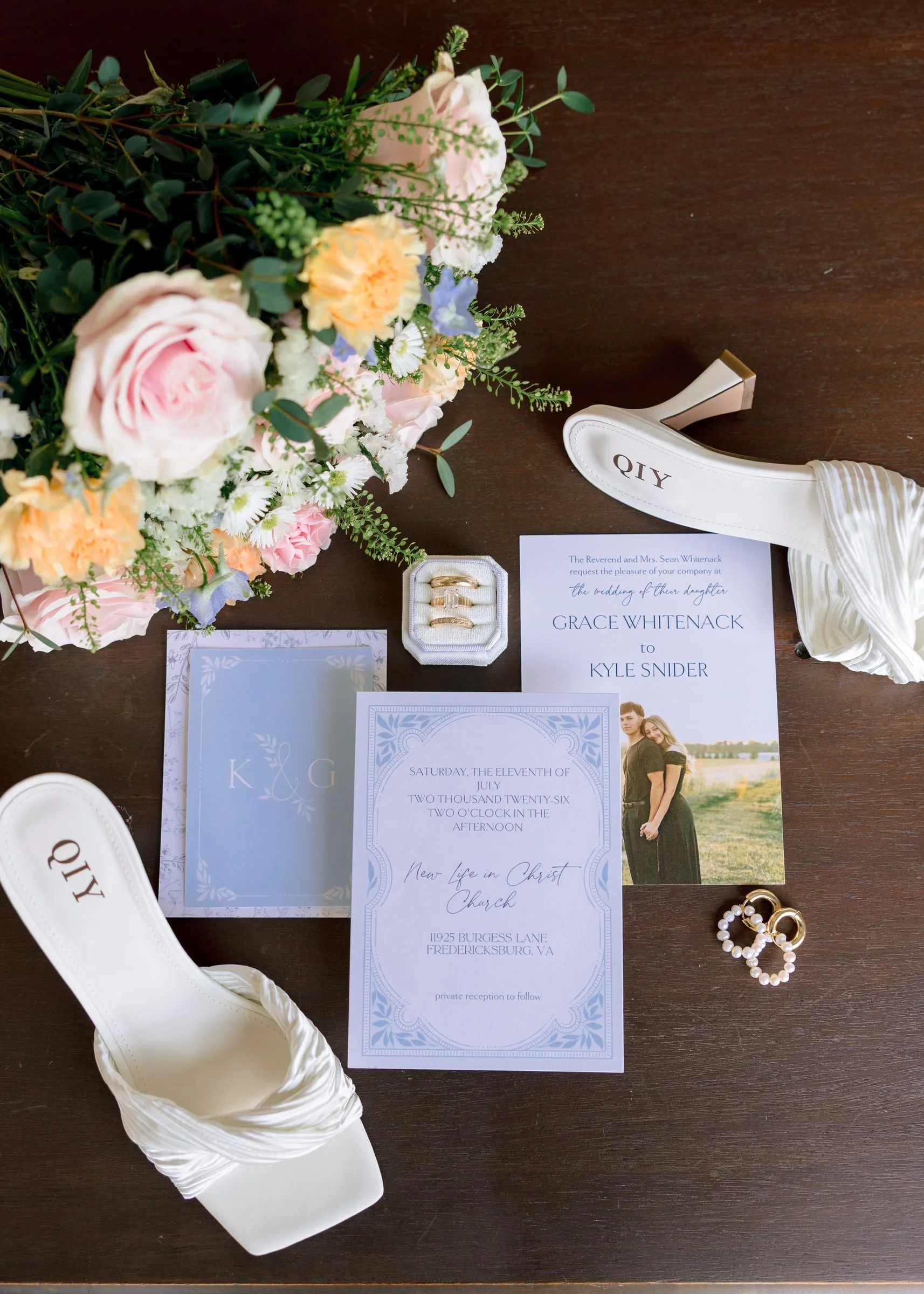 Arrangement of pink, yellow, white, and purple flowers with greenery on a dark wooden surface. Wedding invitation and RSVP cards, white high heel shoes with "QTY" printed on the sole, pearl earrings, and wedding rings.