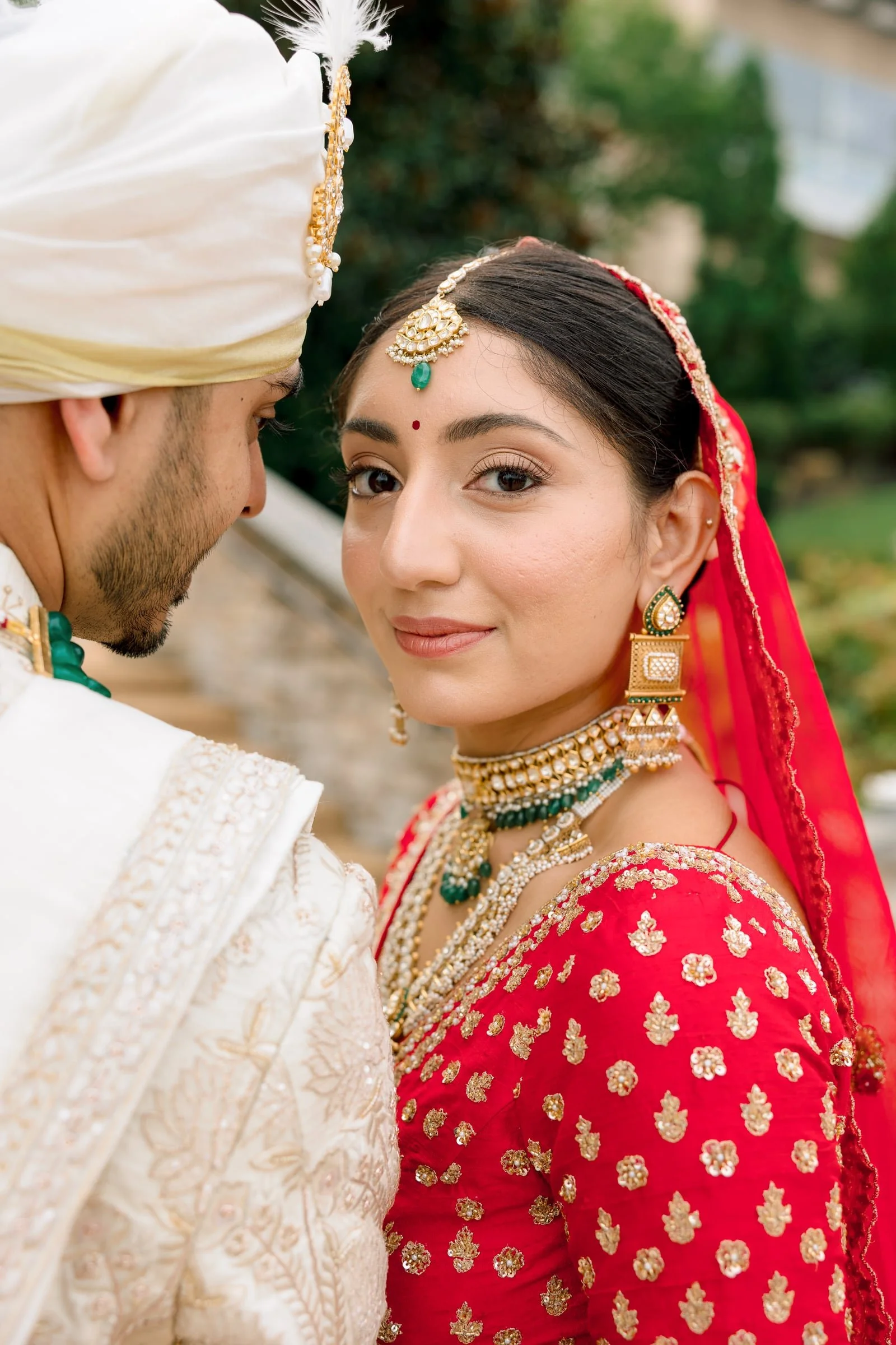 Close-up of a bride and groom in traditional Indian wedding attire, facing each other outdoors.