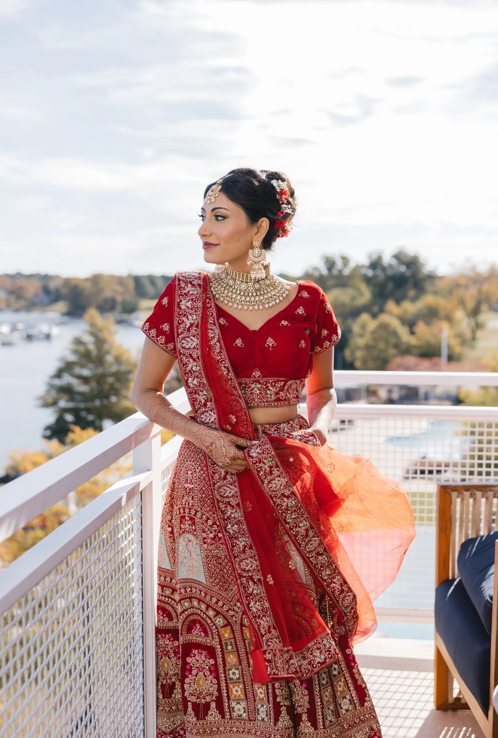 A woman dressed in traditional Indian red bridal attire with intricate gold jewelry, standing on a balcony with an outdoor view of trees and water in the background.