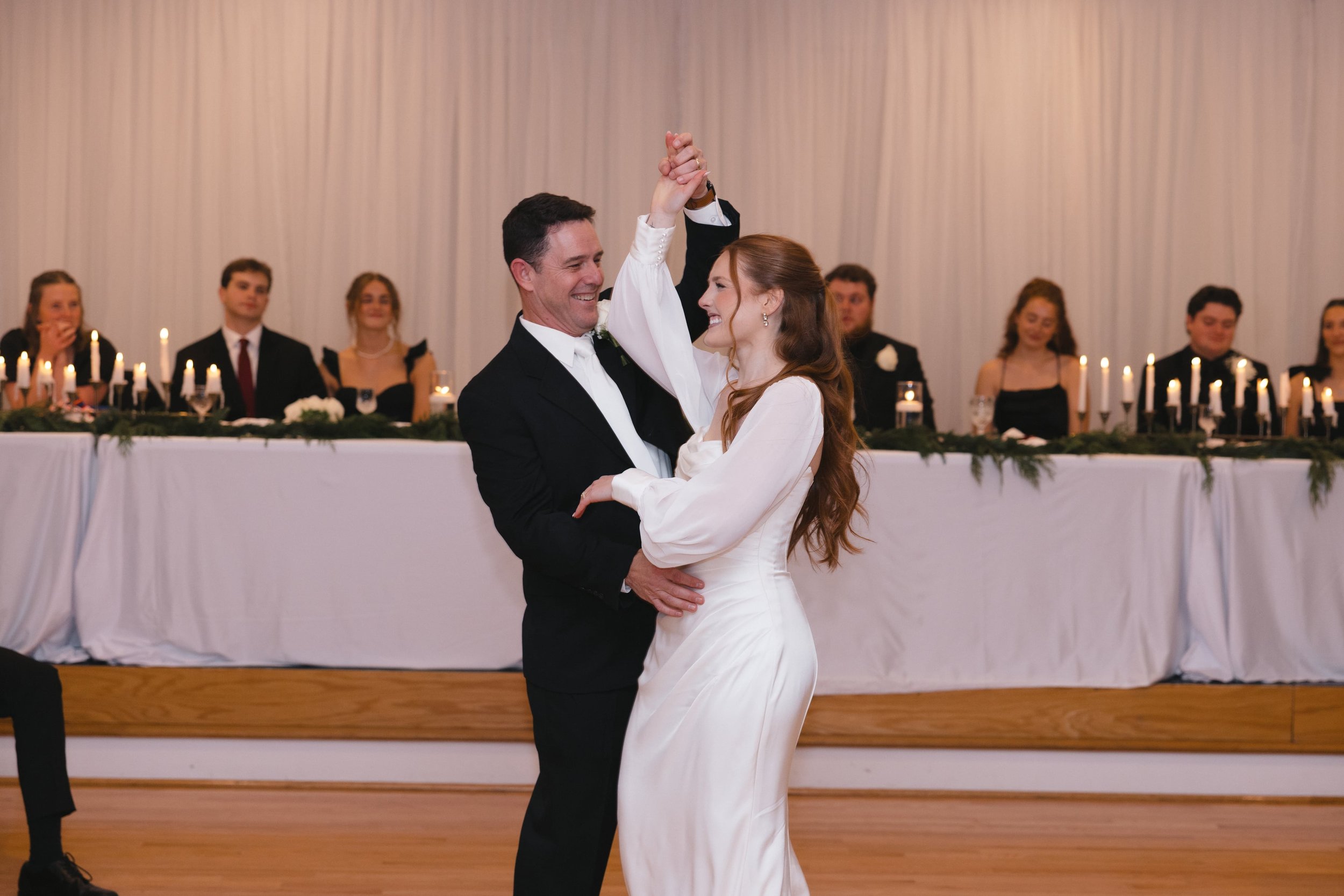 A bride and groom dancing at a wedding reception, with guests seated at a long decorated table in the background.