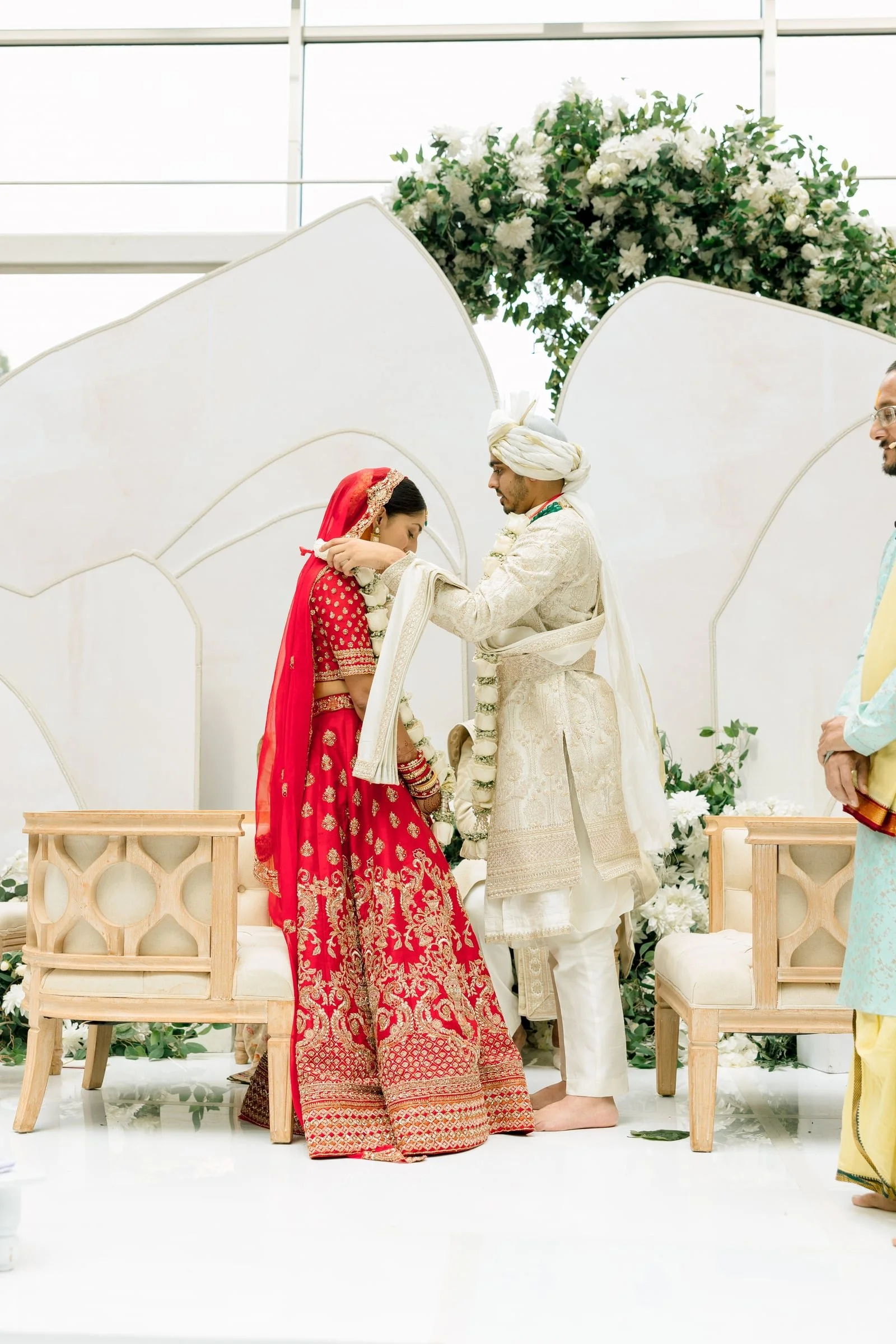 A traditional Indian wedding ceremony with a bride in a red and gold saree and groom in a cream sherwani. They are exchanging vows while standing under a floral arch with white flowers, with a white backdrop and wooden benches.