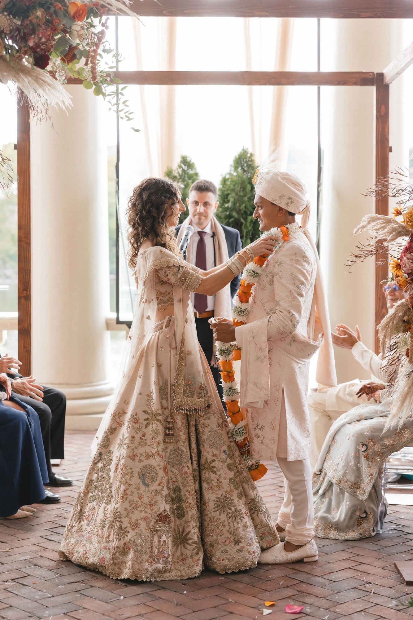 A wedding ceremony with a bride and groom in traditional Indian attire, exchanging garlands in front of an officiant, with guests seated on either side. The bride is in a decorated cream-colored lehenga, and the groom is in a cream sherwani with a tu