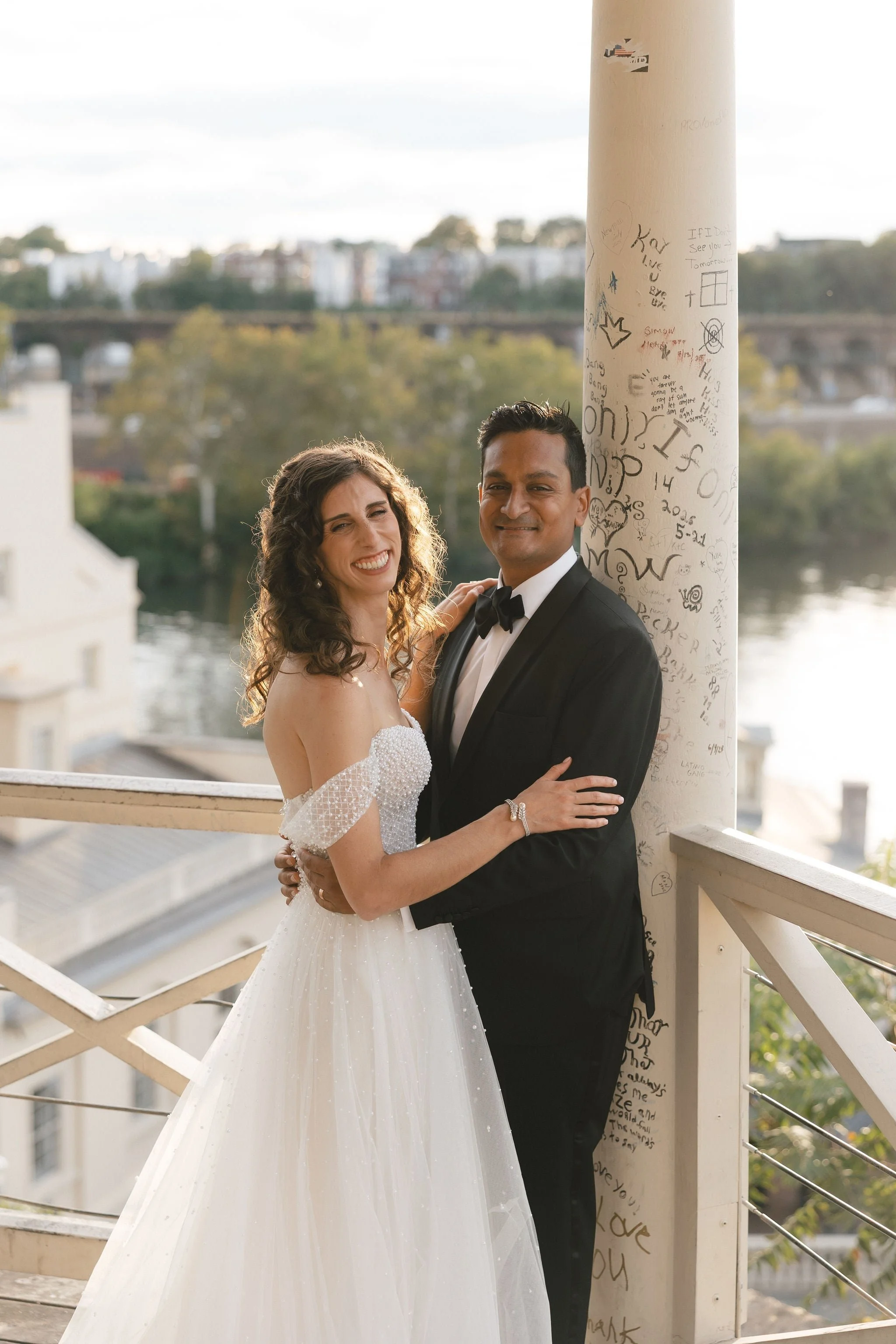 A smiling bride and groom in wedding attire embracing each other on a balcony with graffiti-covered white pillar, cityscape, and river in the background.