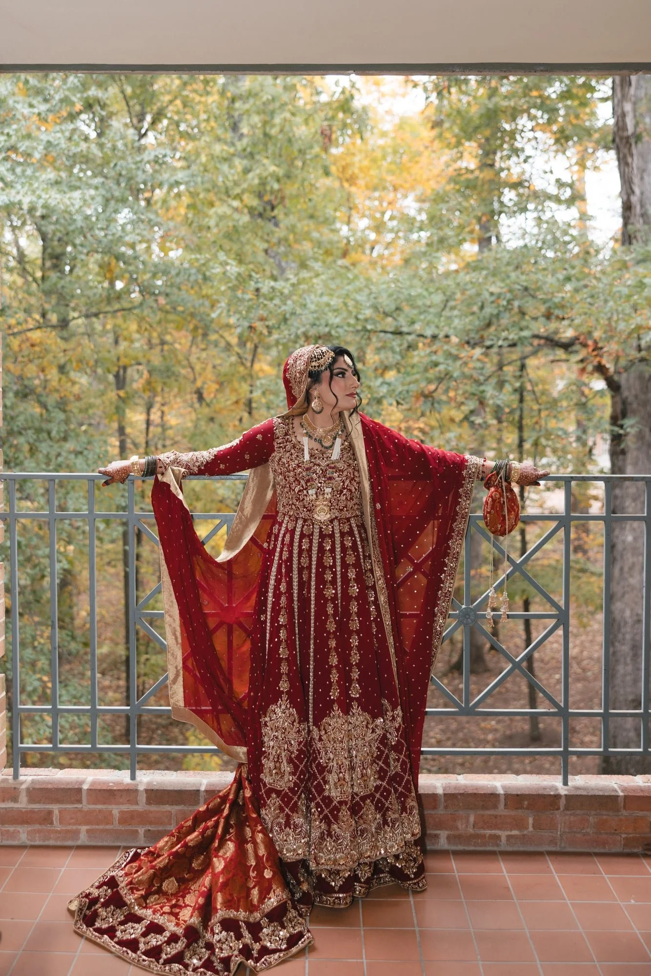A woman wearing a traditional red and gold ornate wedding dress with intricate embroidery and jewelry, standing upscale on a balcony with a metal railing, surrounded by autumn foliage trees.