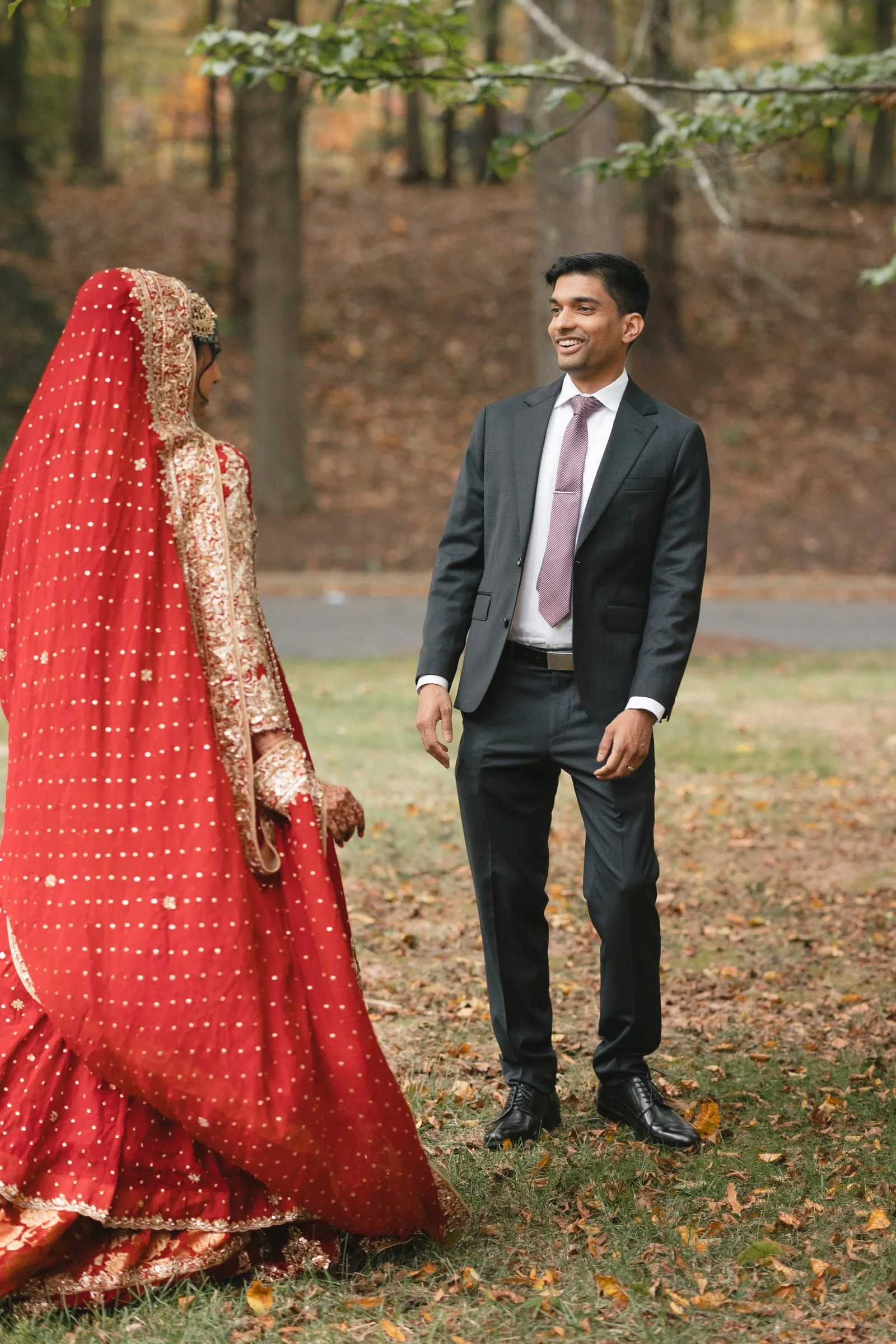 A man and woman dressed in formal attire are standing outdoors in a wooded area with fallen autumn leaves. The woman, on the left, is wearing a traditional red Indian bridal outfit with gold embroidery and a matching dupatta covering her head. The ma