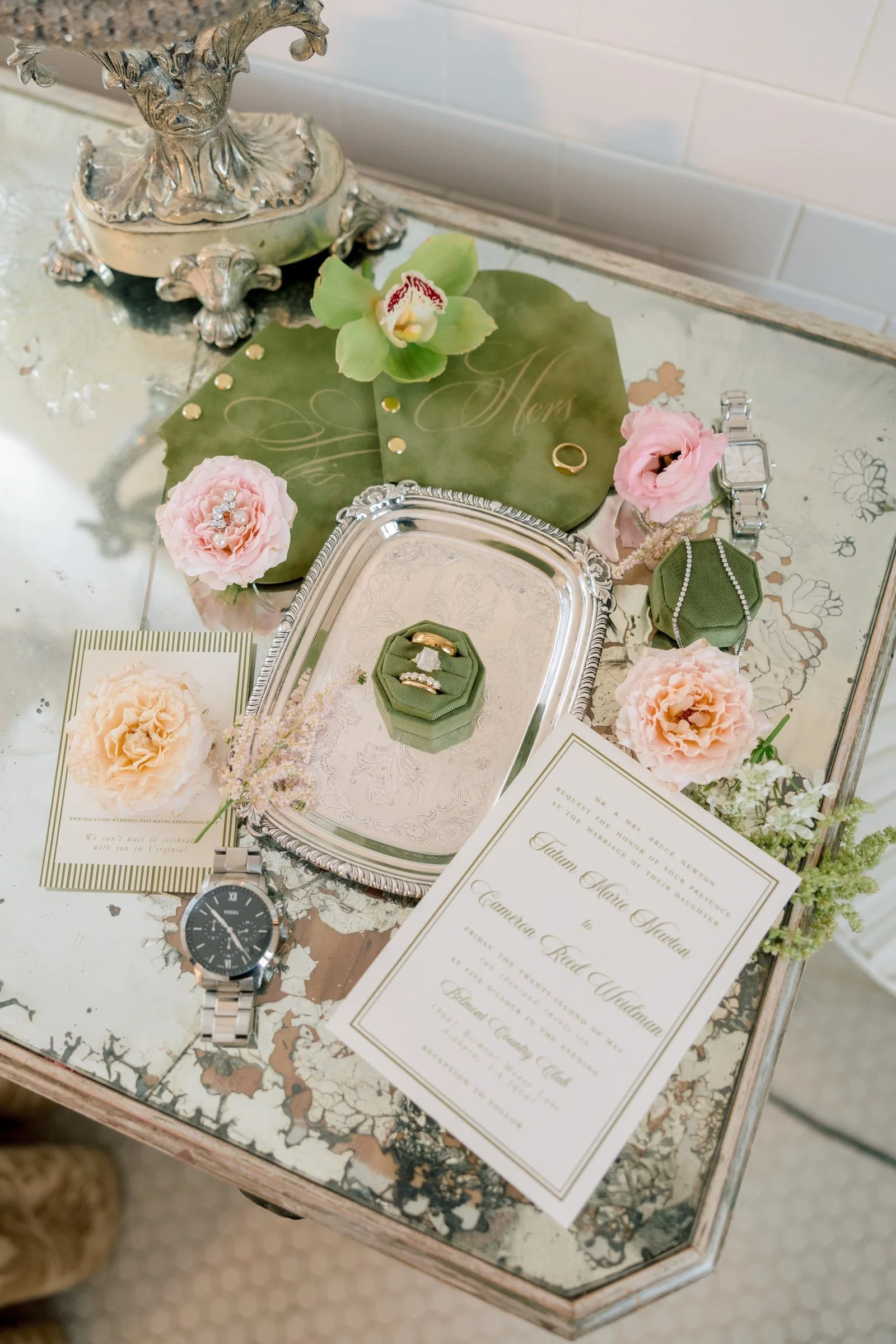 An ornate silver tray on a distressed table with a wedding invitation, a wristwatch, a ring, a jewelry box, and decorative flowers, including pink and peach blossoms and a green orchid, signifying a wedding setting.