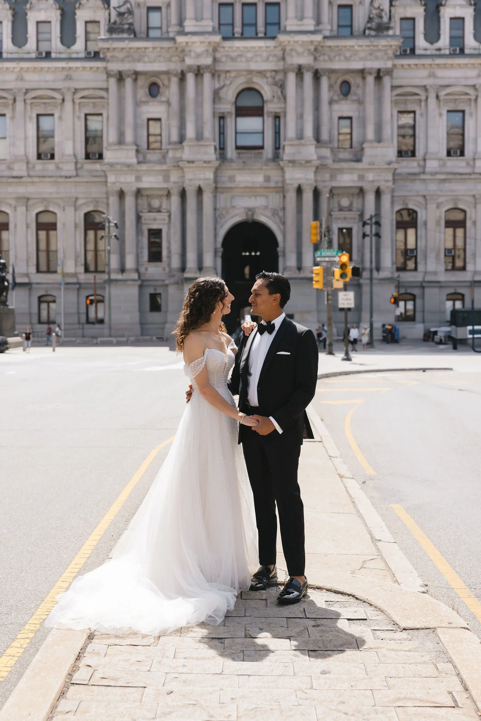 A bride and groom standing on a city street in front of a historic building, smiling and holding hands, dressed in wedding attire.
