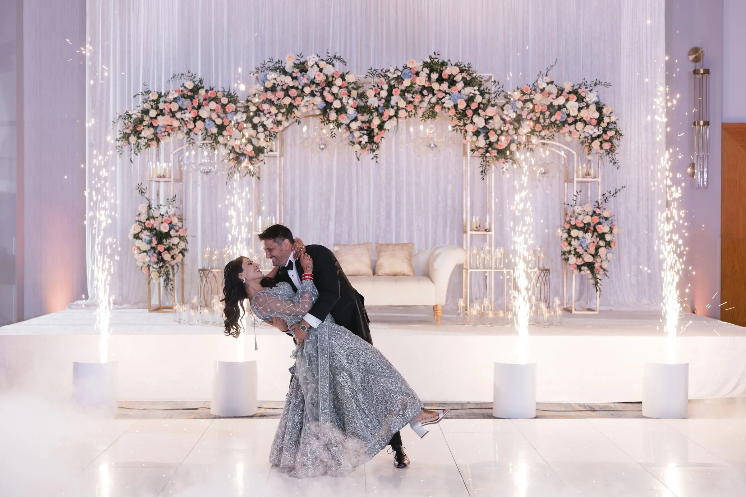 A bride and groom dancing on a wedding stage decorated with a floral arch, surrounded by sparklers.