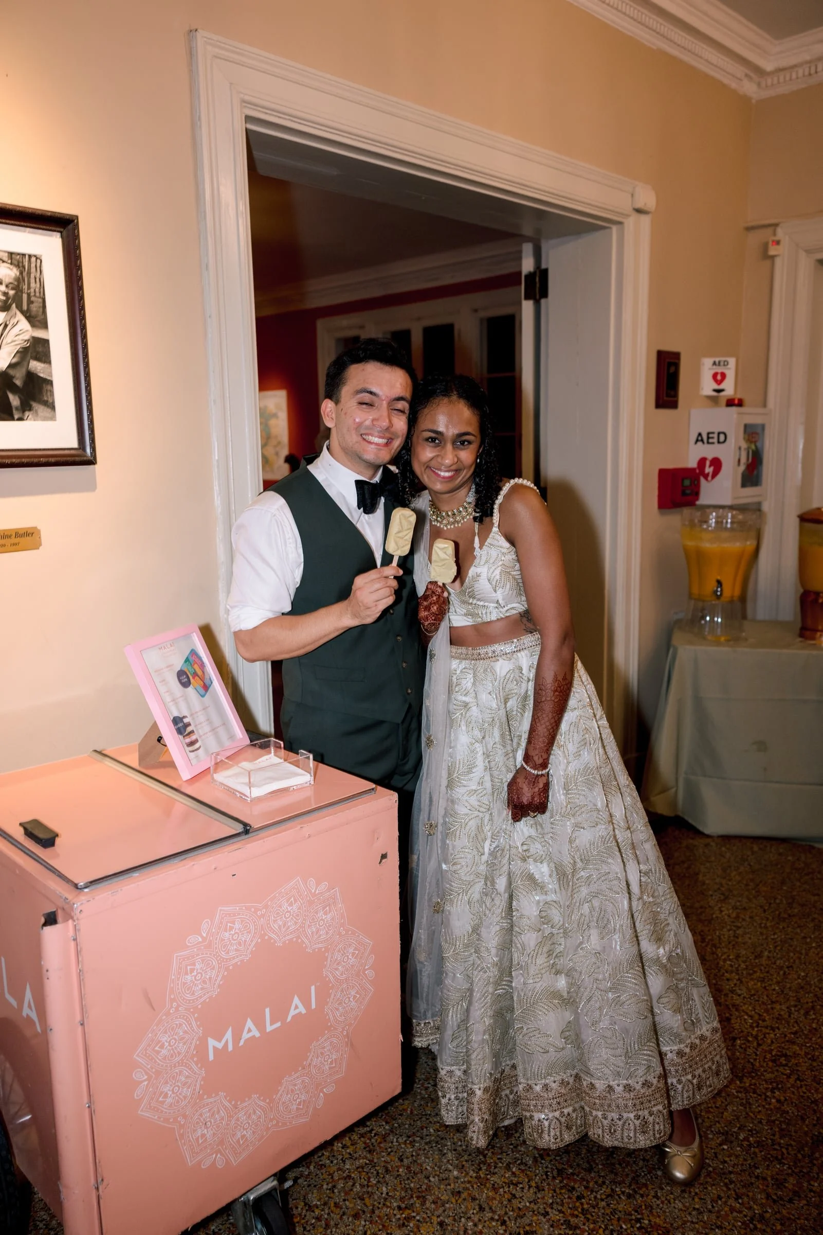 A couple in wedding attire smiling and holding ice cream bars, standing inside a building near a pink dessert cart with the word 'Malai' on it. The man is wearing a black vest, white shirt, and black bow tie, and the woman is wearing a white bridal g