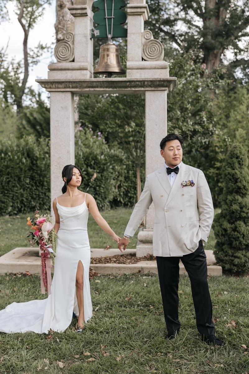 A bride and groom holding hands outdoors, standing in front of a stone arch with a bell. The bride wears a white dress with a slit, and the groom wears a light-colored tuxedo with a black bow tie. They are surrounded by trees and greenery.