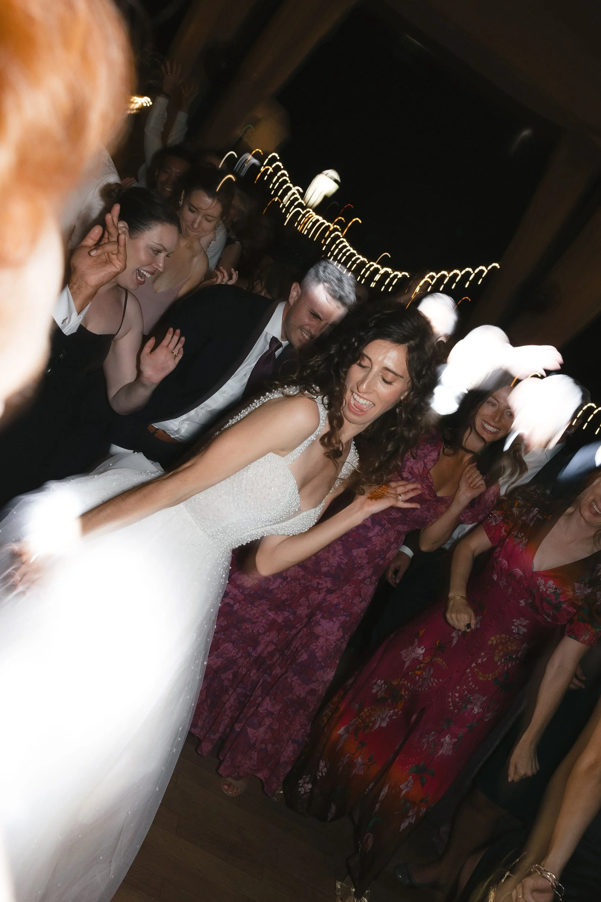 People dancing and celebrating at a wedding reception, with a woman in a white wedding dress in the foreground and a group of women in red and purple dresses behind her, all smiling and enjoying themselves.
