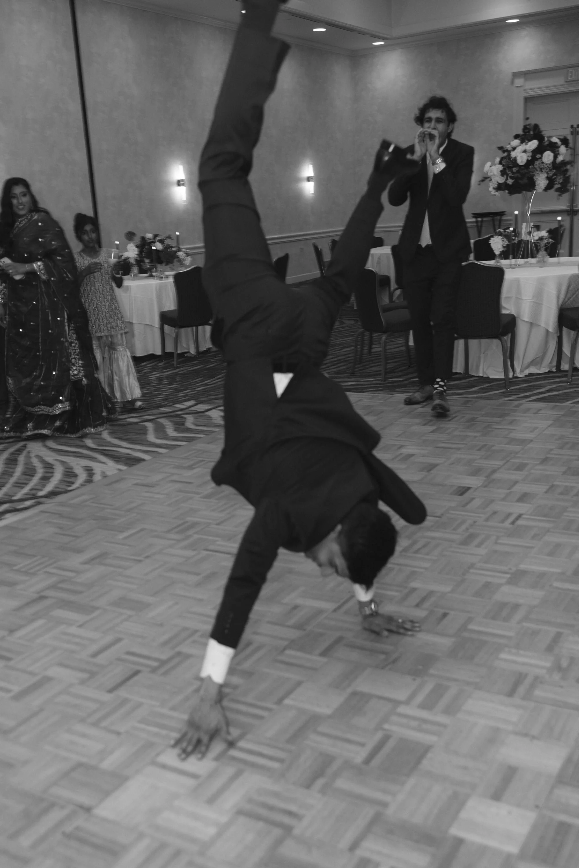 Man performing a break dance move on a dance floor in a formal setting, with a woman and children observing, and tables with floral arrangements in the background.
