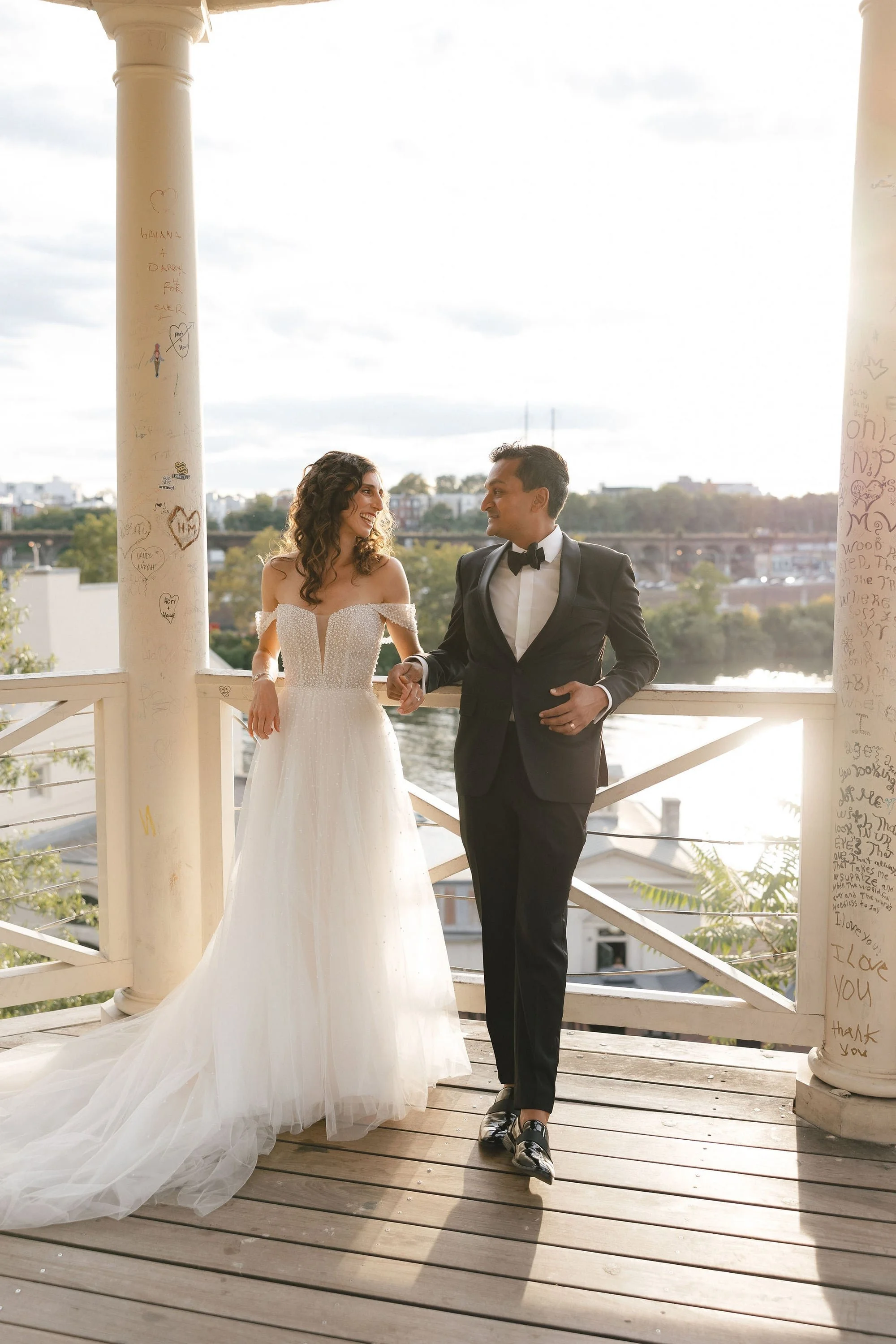 A bride and groom holding hands and smiling at each other on a wooden balcony with graffiti-covered columns, overlooking a cityscape with water and trees in the background, during sunset.