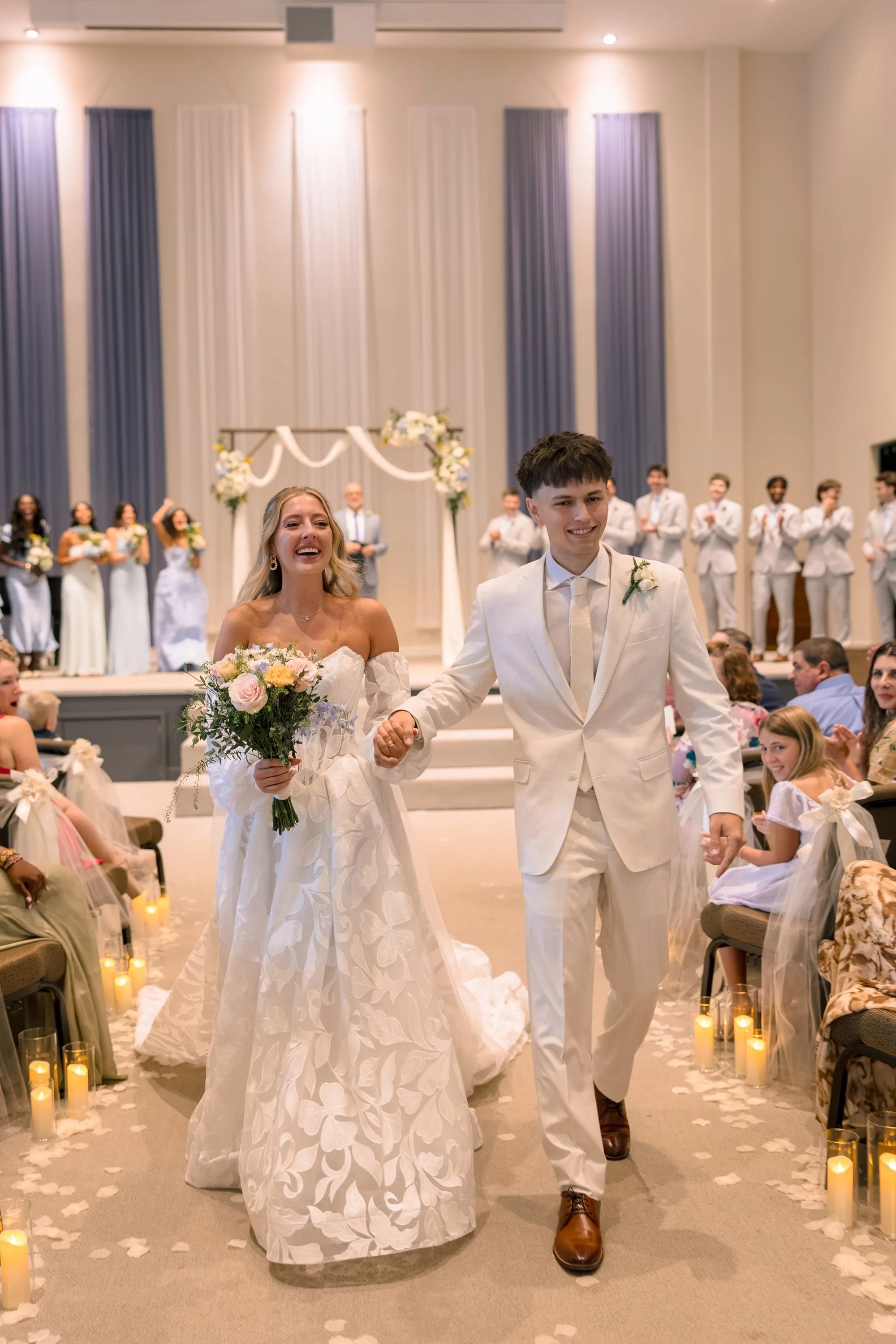 A bride and groom holding hands walking down the aisle at their wedding ceremony with guests watching, candles on the floor, and bridesmaids and groomsmen in the background.
