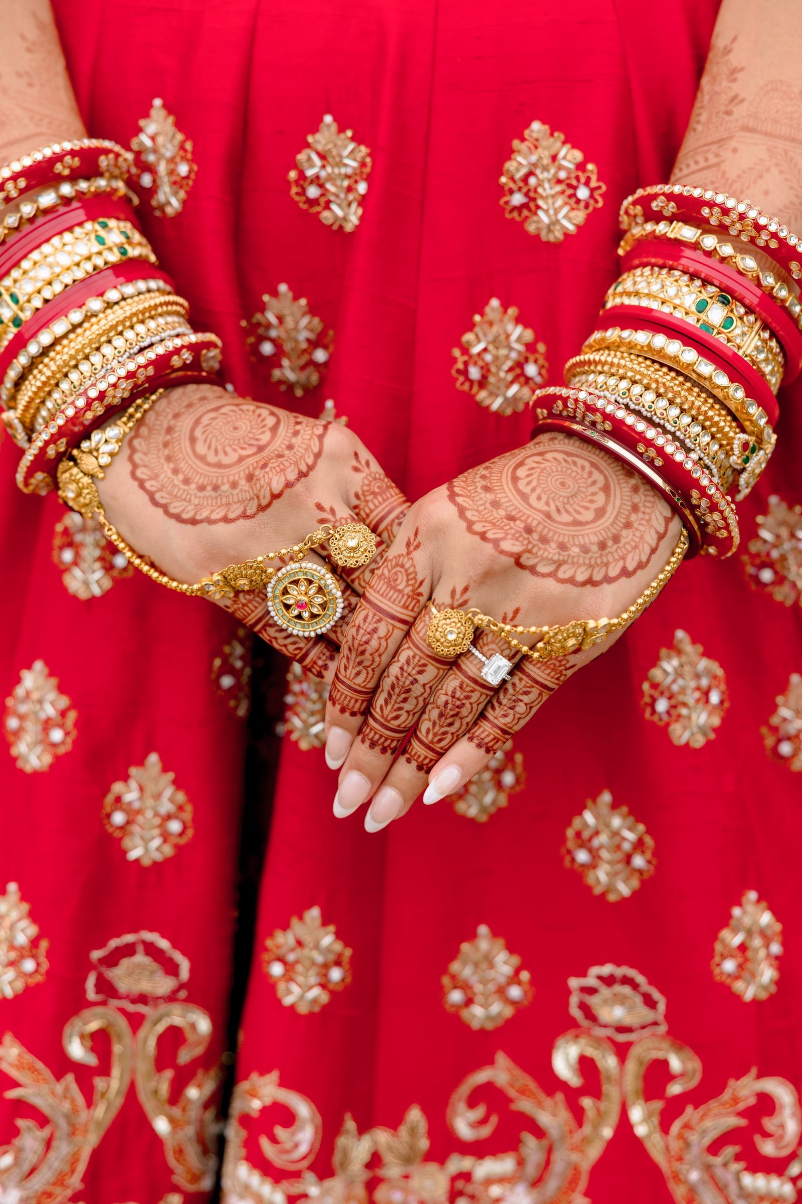 Close-up of woman's hands adorned with henna designs and gold jewelry, wearing a red traditional outfit with gold embroidery and embellishments.