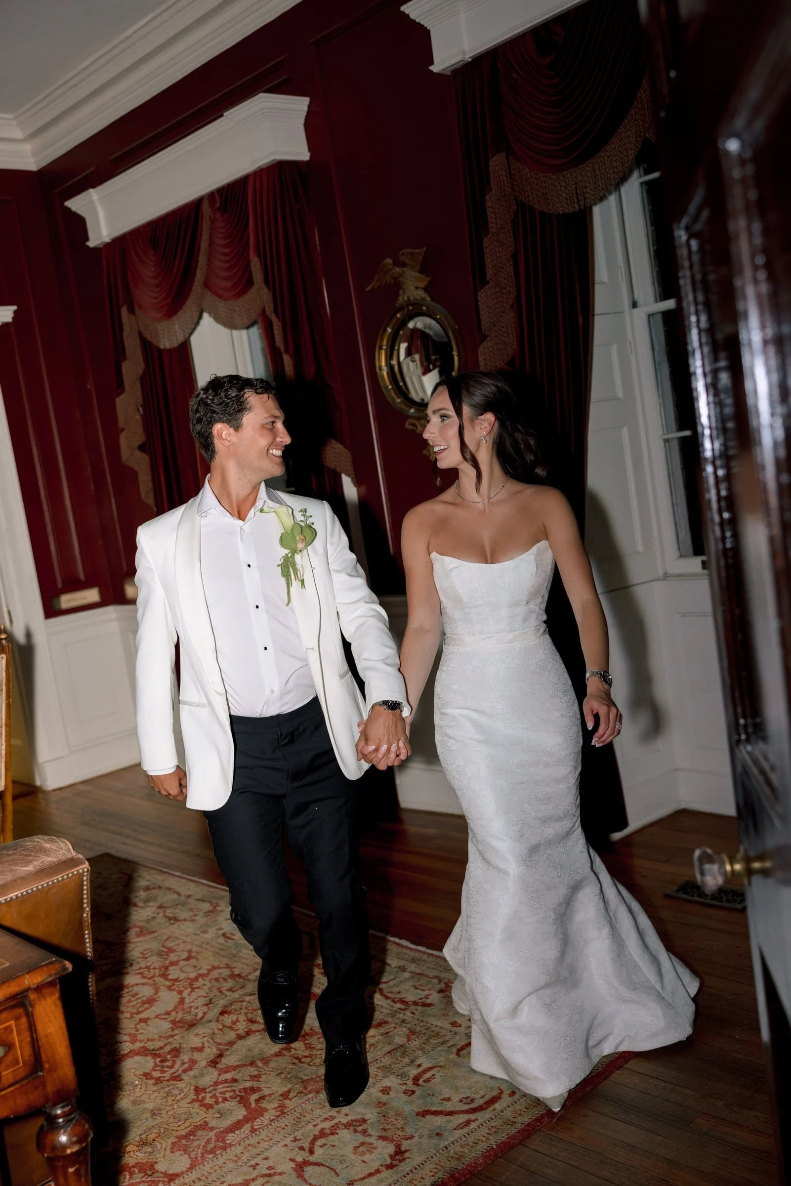 A bride and groom holding hands and smiling at each other inside a well-lit room with elegant dark reds and white decorations. The groom is wearing a white tuxedo jacket with a green boutonniere, while the bride is wearing a strapless white wedding d