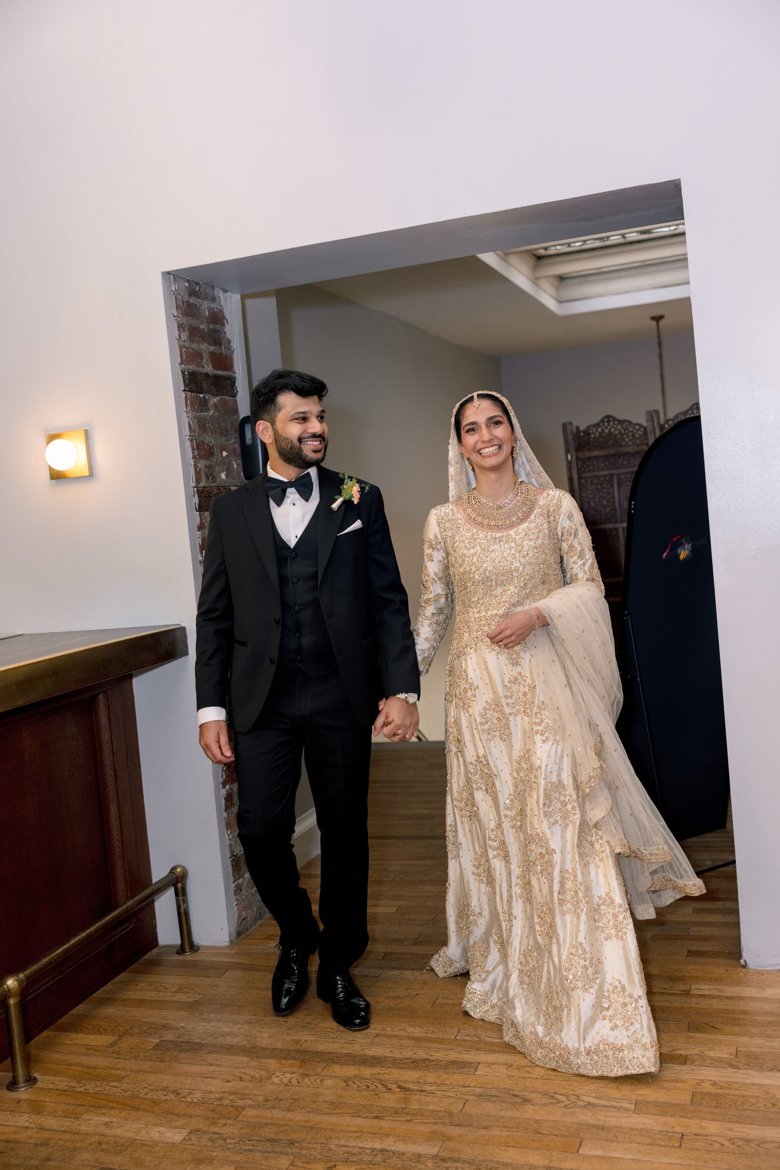 A happy couple dressed in formal wedding attire, holding hands and smiling, standing in a room with wooden flooring and white walls, with a decorative screened room visible in the background.