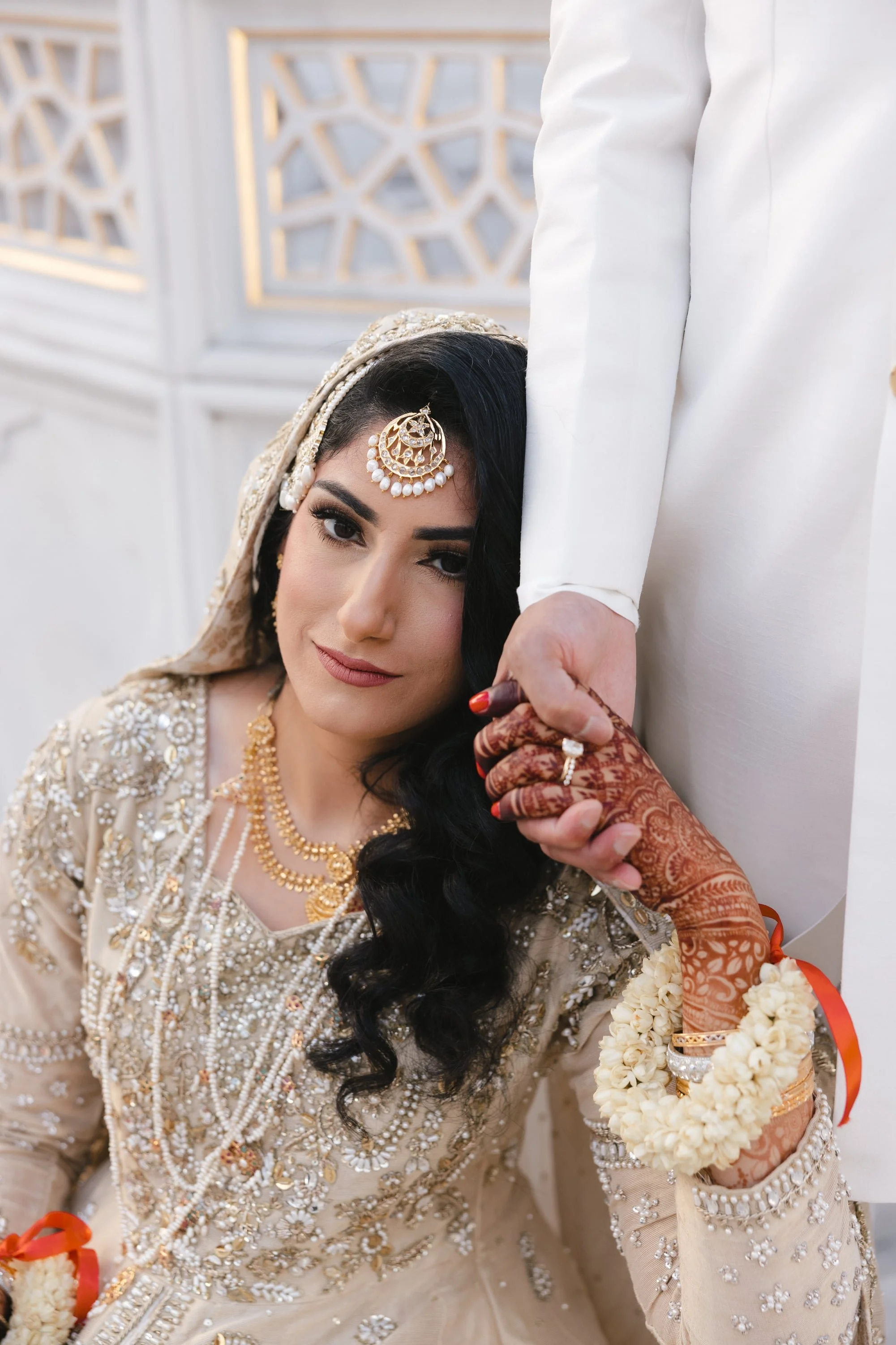 A woman dressed in traditional Indian bridal attire, with intricate jewelry and henna on her hand, leaning her head against a man's hand.