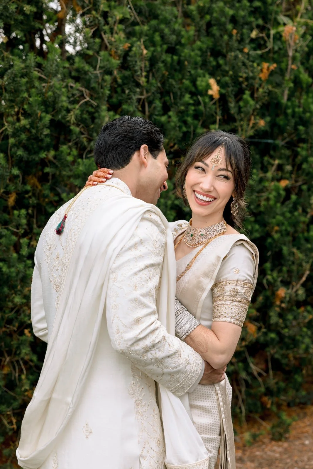 A joyful couple dressed in traditional Indian wedding attire, embracing outdoors with greenery in the background.
