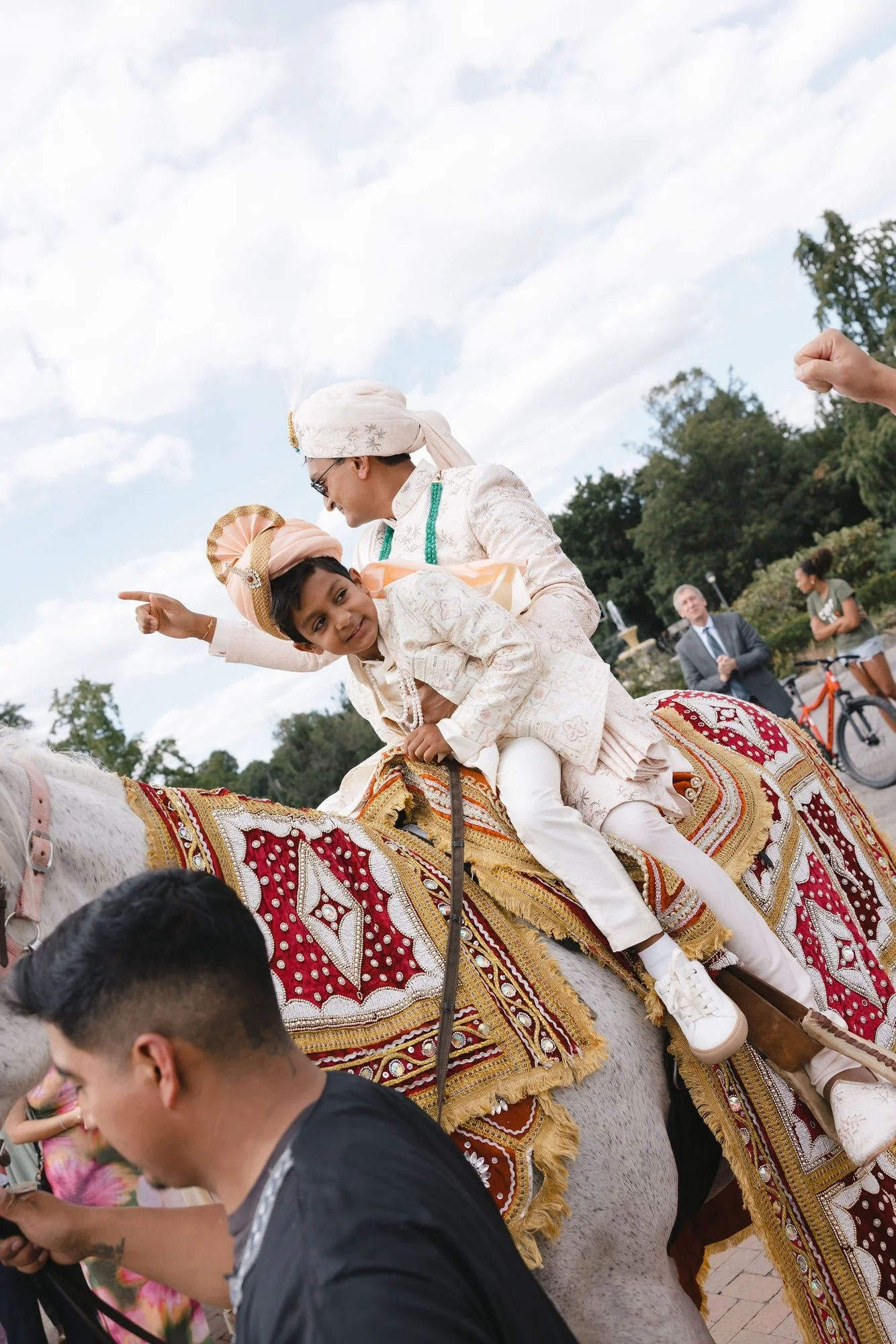 People riding a decorated white horse during a cultural celebration, with onlookers and a cloudy sky in the background.