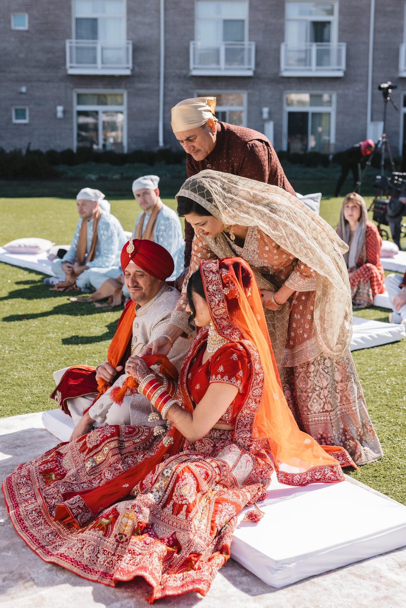 People dressed in traditional Indian wedding attire, seated on a white platform outdoors during a cultural ceremony.