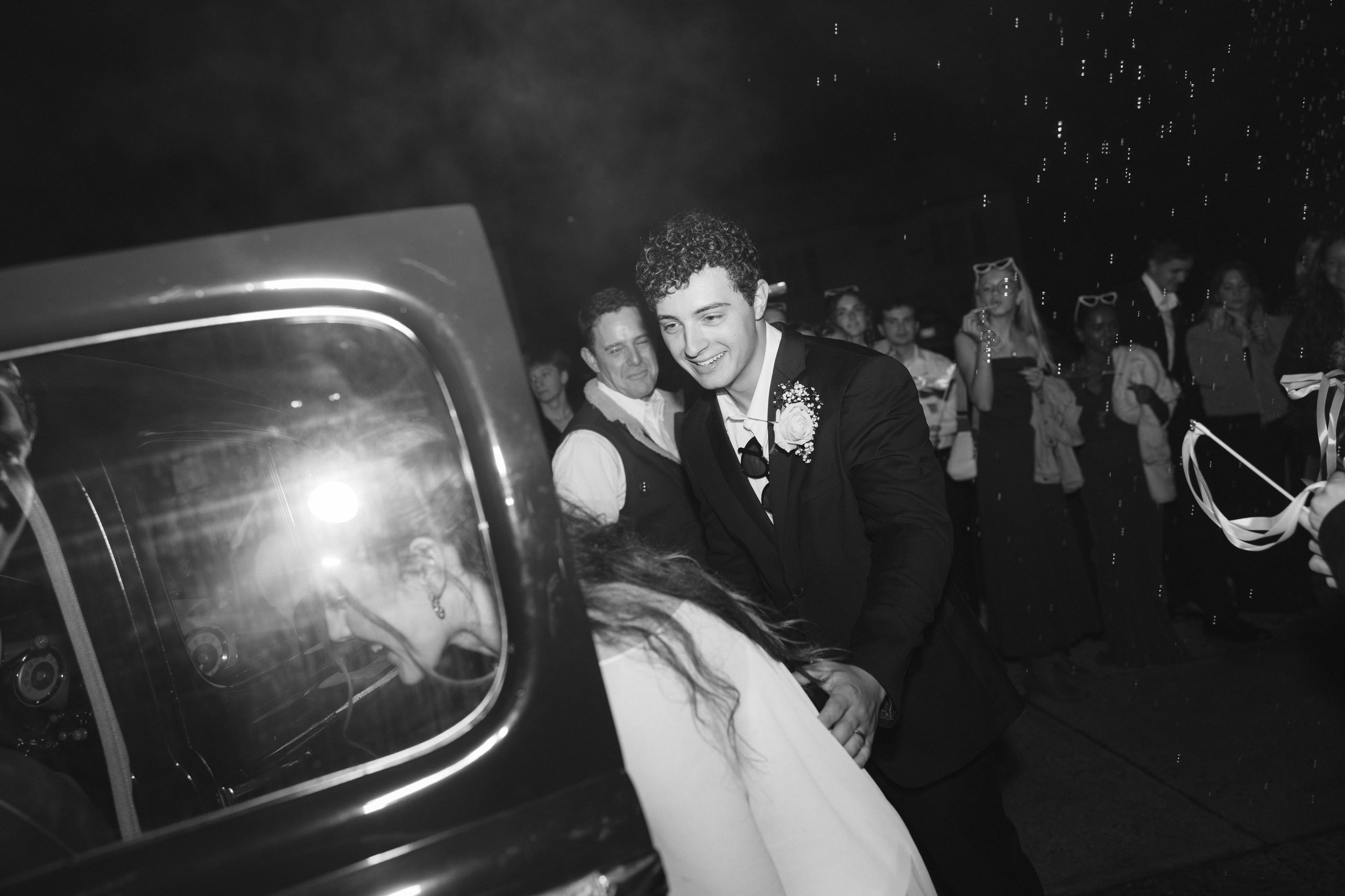 A young man in a tuxedo with a boutonnière smiling and interacting with guests at a celebration or party, with a group of people in the background.