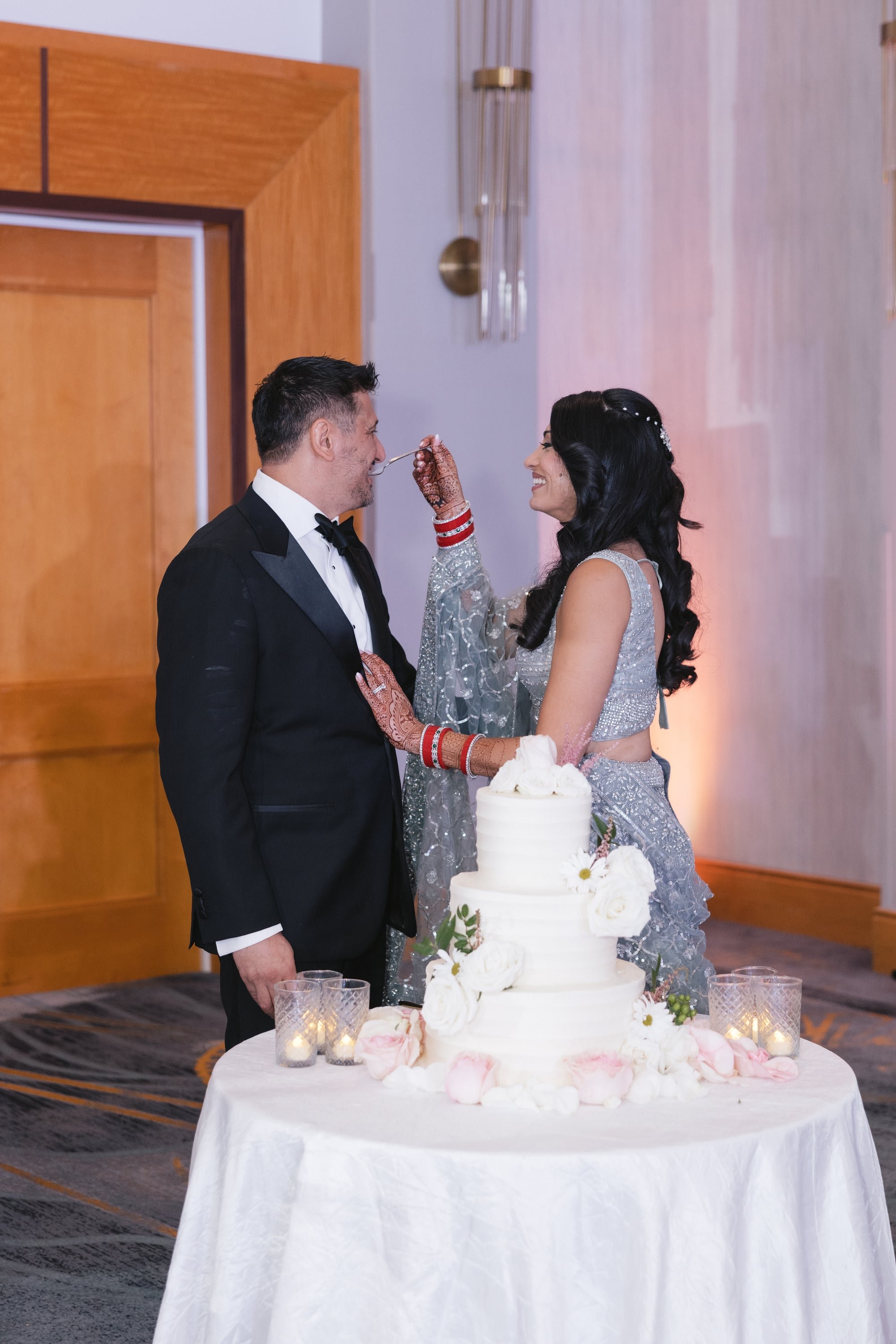 A bride and groom are smiling and feeding each other cake at their wedding reception. They are standing behind a round table with a white tablecloth, decorated with flowers and candles. The bride is dressed in a silver gown with matching hijab and he