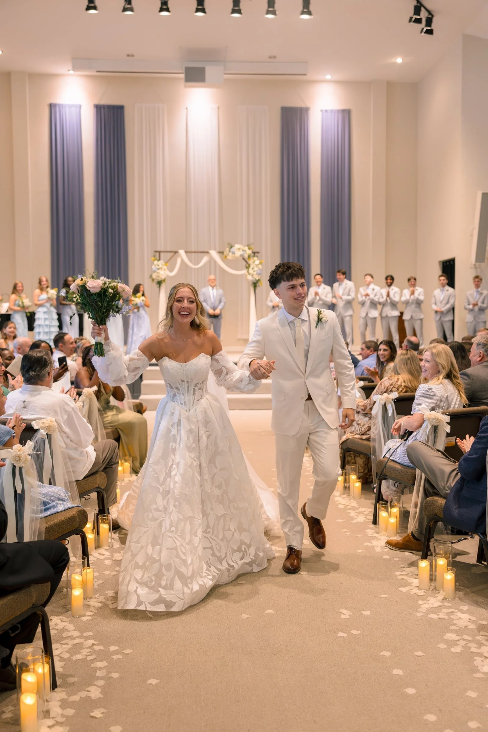A newlywed couple walking down the aisle after their wedding ceremony, surrounded by seated guests in a decorated indoor venue with candles and flower petals on the floor.