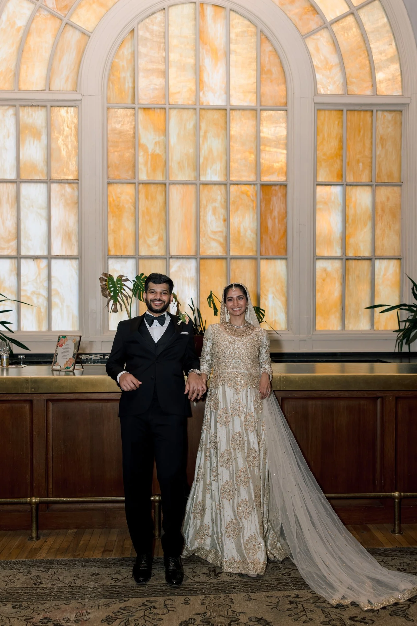A bride and groom standing in front of a large stained glass window with warm tones, inside a historic building. The groom is wearing a tuxedo with a bow tie, and the bride is in an ornate, gold-embellished gown with a veil.