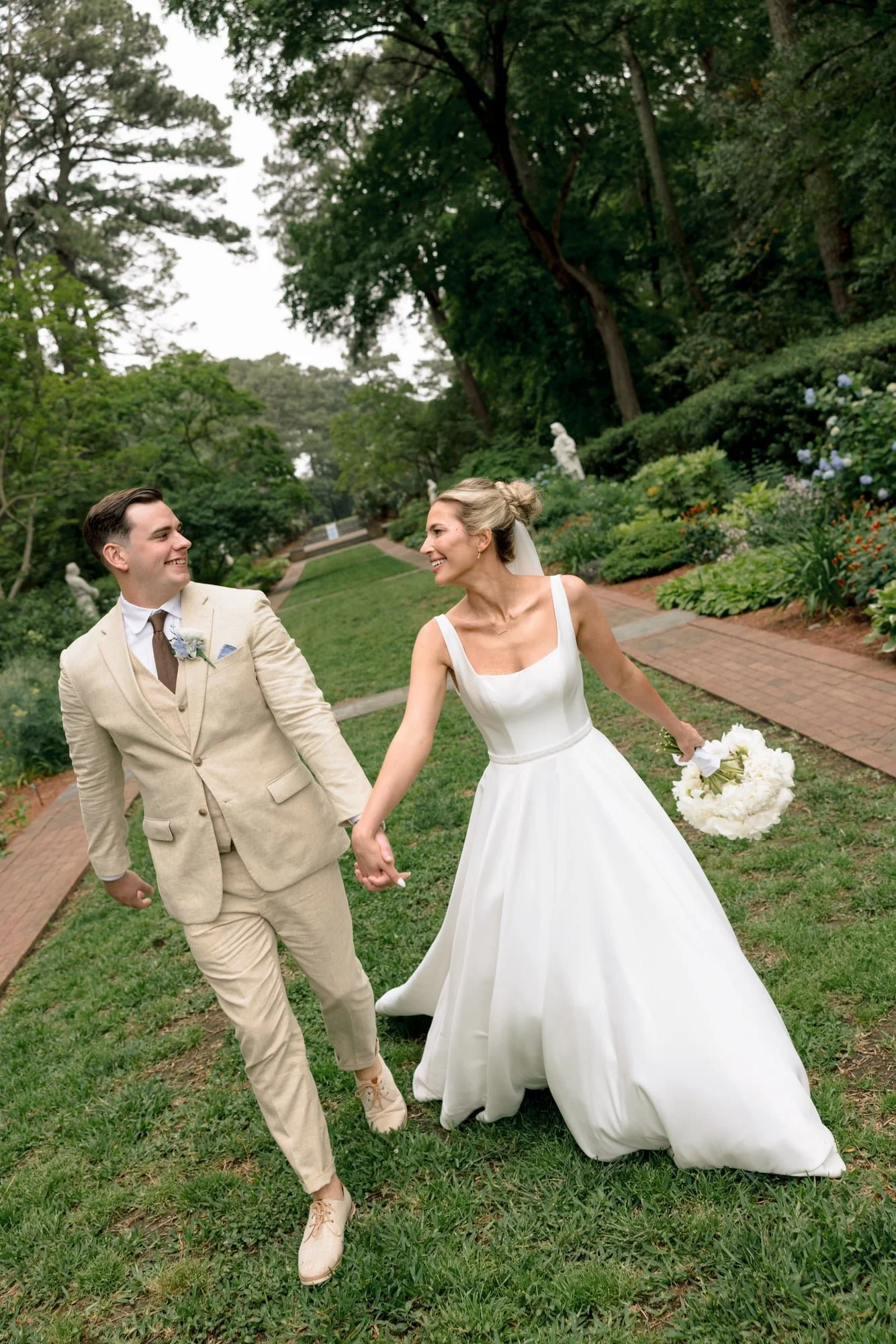 A bride and groom holding hands and smiling as they walk through a lush garden with trees, bushes, and flower beds.