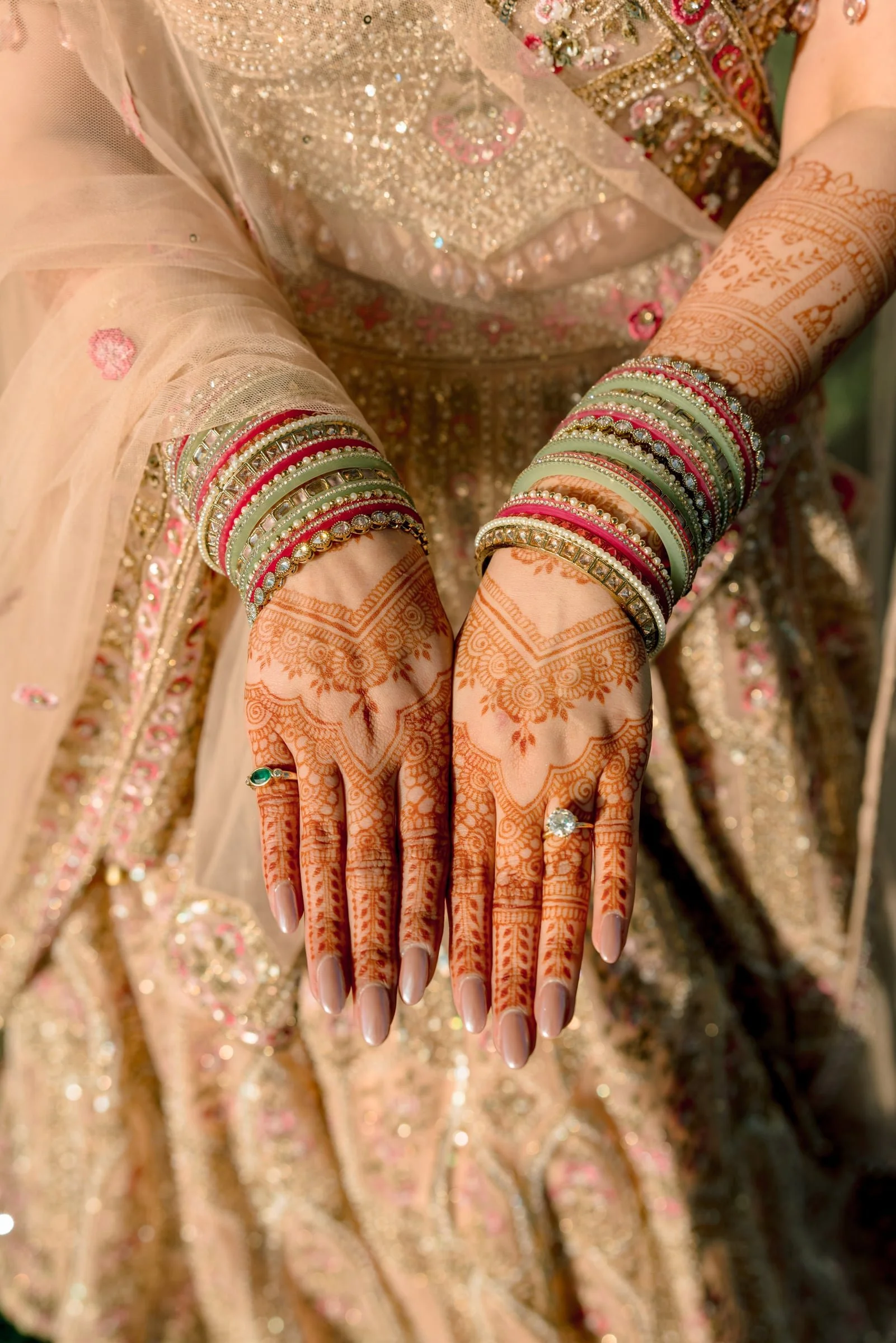 Close-up of a woman’s hands displaying intricate mehndi (henna) designs and wearing numerous colorful bangles, in front of traditional, ornate Indian clothing.
