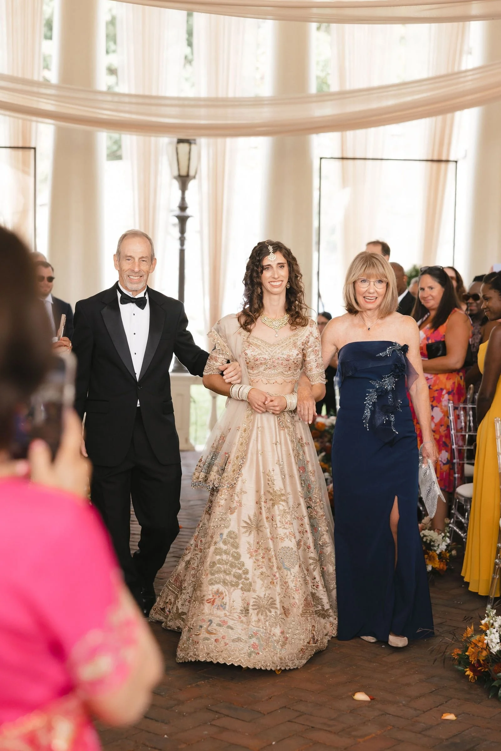 A bride walking down the aisle at her wedding reception, dressed in an ornate traditional Indian bridal dress with jewelry, flanked by two older individuals, with wedding guests in the background.